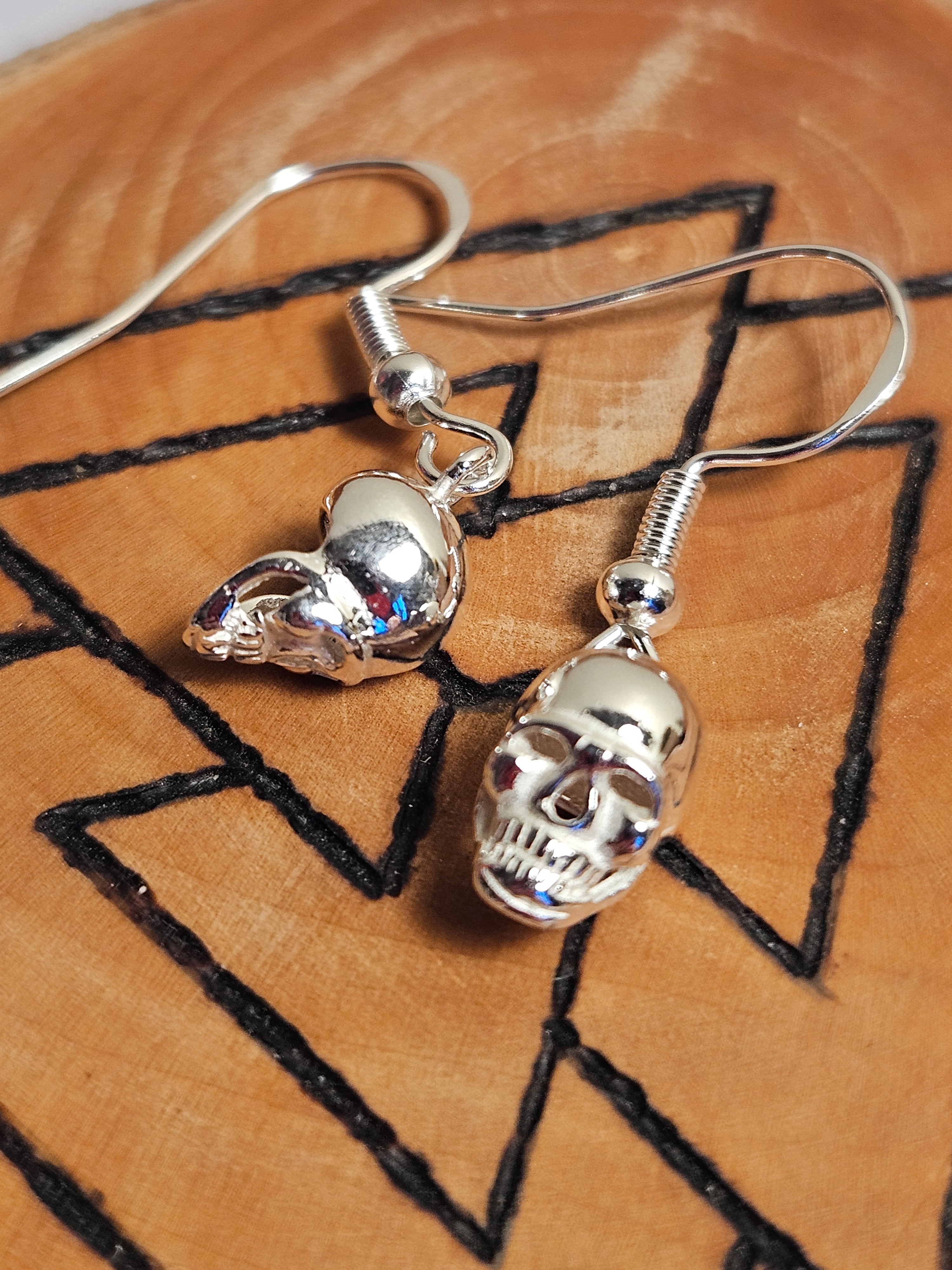 Silver skull earrings on a wooden surface with black line art.