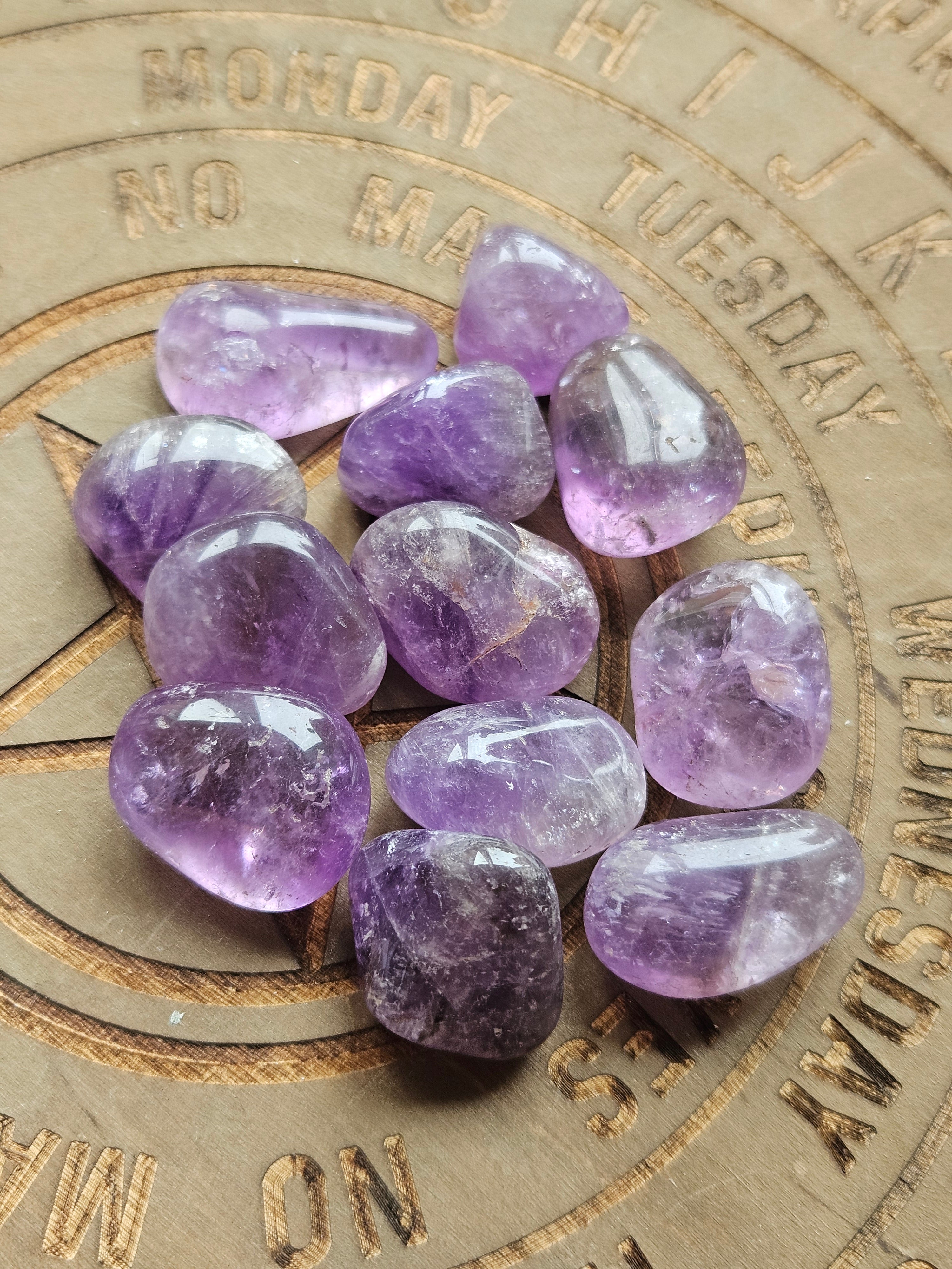 A group of polished purple amethyst tumbled stones arranged on a circular wooden board with engraved days of the week.