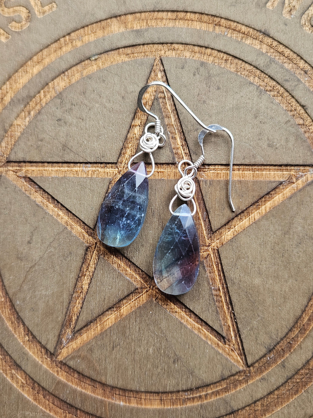 A pair of teardrop-shaped dangle earrings with iridescent blue and purple Fluorite stones in silver wire-wrapped hooks, displayed on a wooden pentagram board.