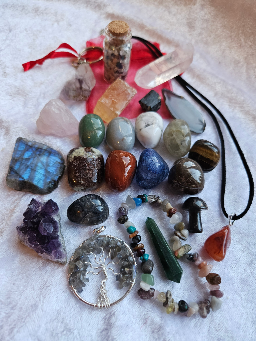 Advent calendar contents, a collection of colorful raw gemstones, polished stones, crystals, and jewelry pieces arranged on a white textured fabric background. The assortment includes a tree of life pendant, a purple crystal cluster, a green crystal pendant, a crystal chip bracelet, and various other raw stones and a small vial with a cork stopper.