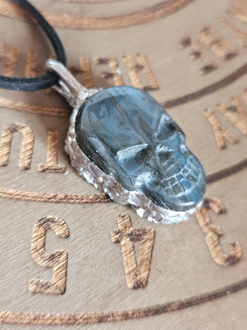 A skull-shaped pendant made of dark gray stone with a textured silver border, hanging from a black cord, set against a beige background with engraved markings.