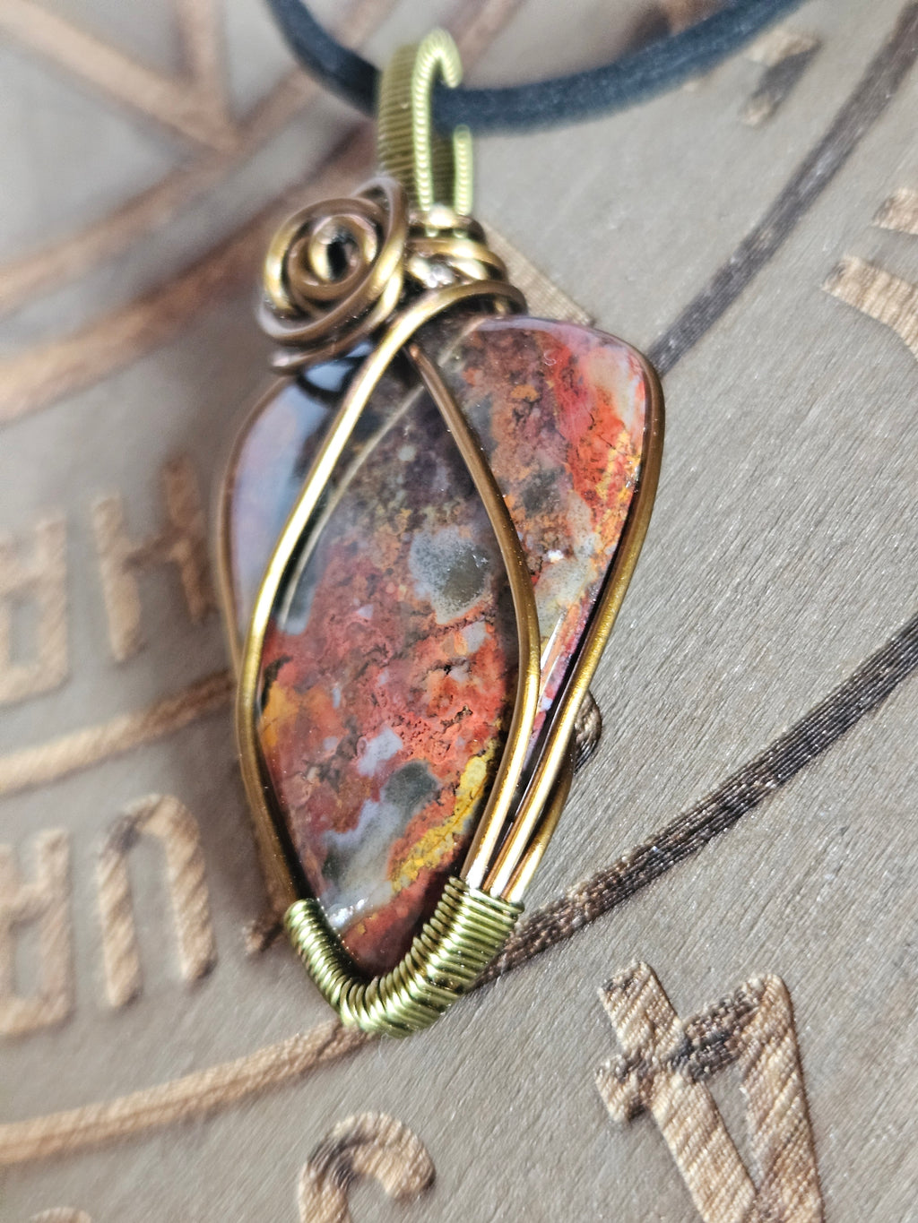 A close-up of a handmade wire-wrapped pendant featuring a textured, mottled stone with orange, brown, and gray tones, set in an antique gold-toned metal frame with a dark blue cord.