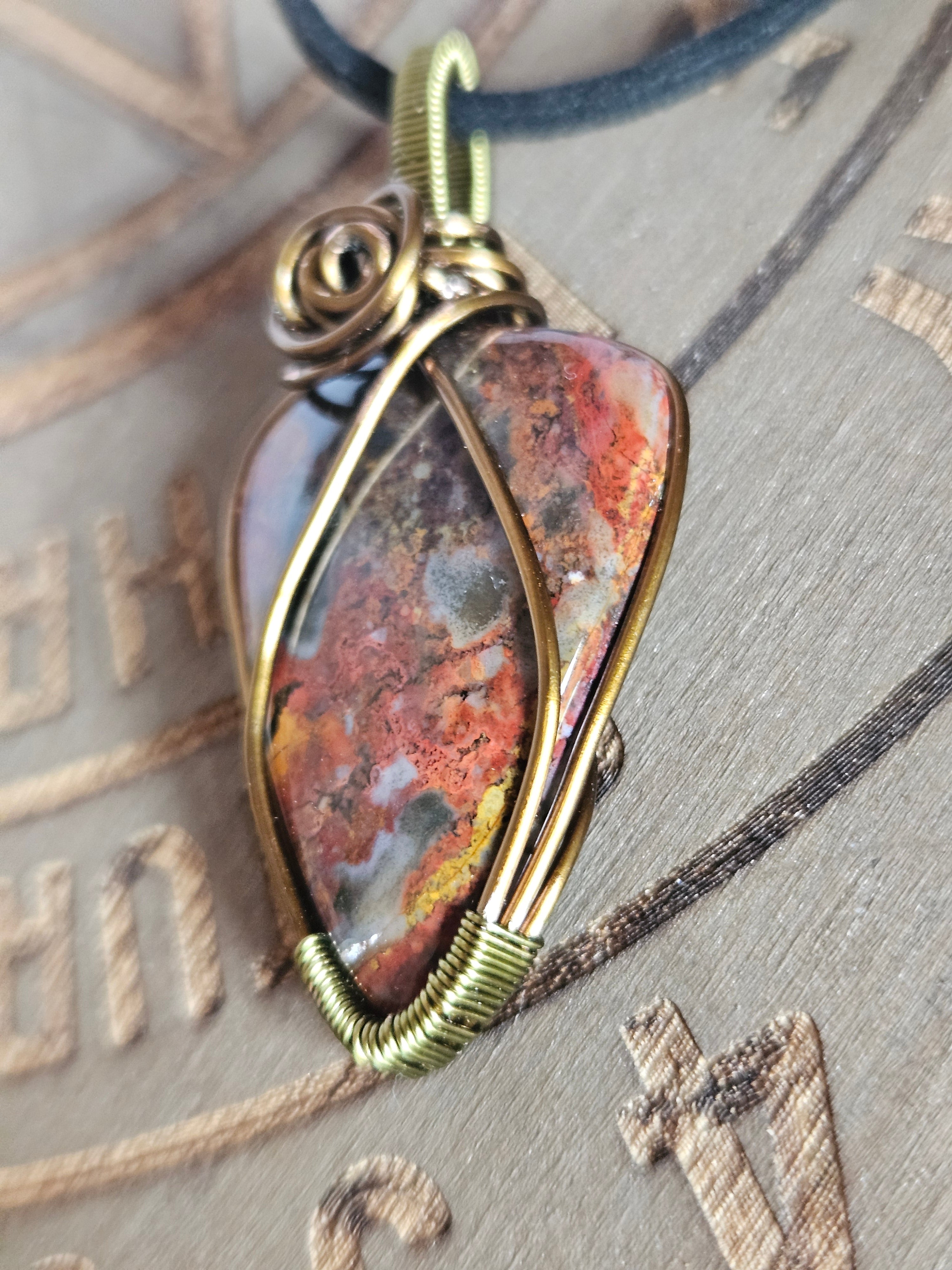 A close-up of a handmade wire-wrapped pendant featuring a textured, mottled stone with orange, brown, and gray tones, set in an antique gold-toned metal frame with a dark blue cord.