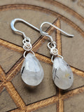 A pair of teardrop-shaped dangle earrings made from Tourmaline In Quartz, faceted gemstone drops in silver wire-wrapping and fishhook ear wires, displayed on a wooden background.