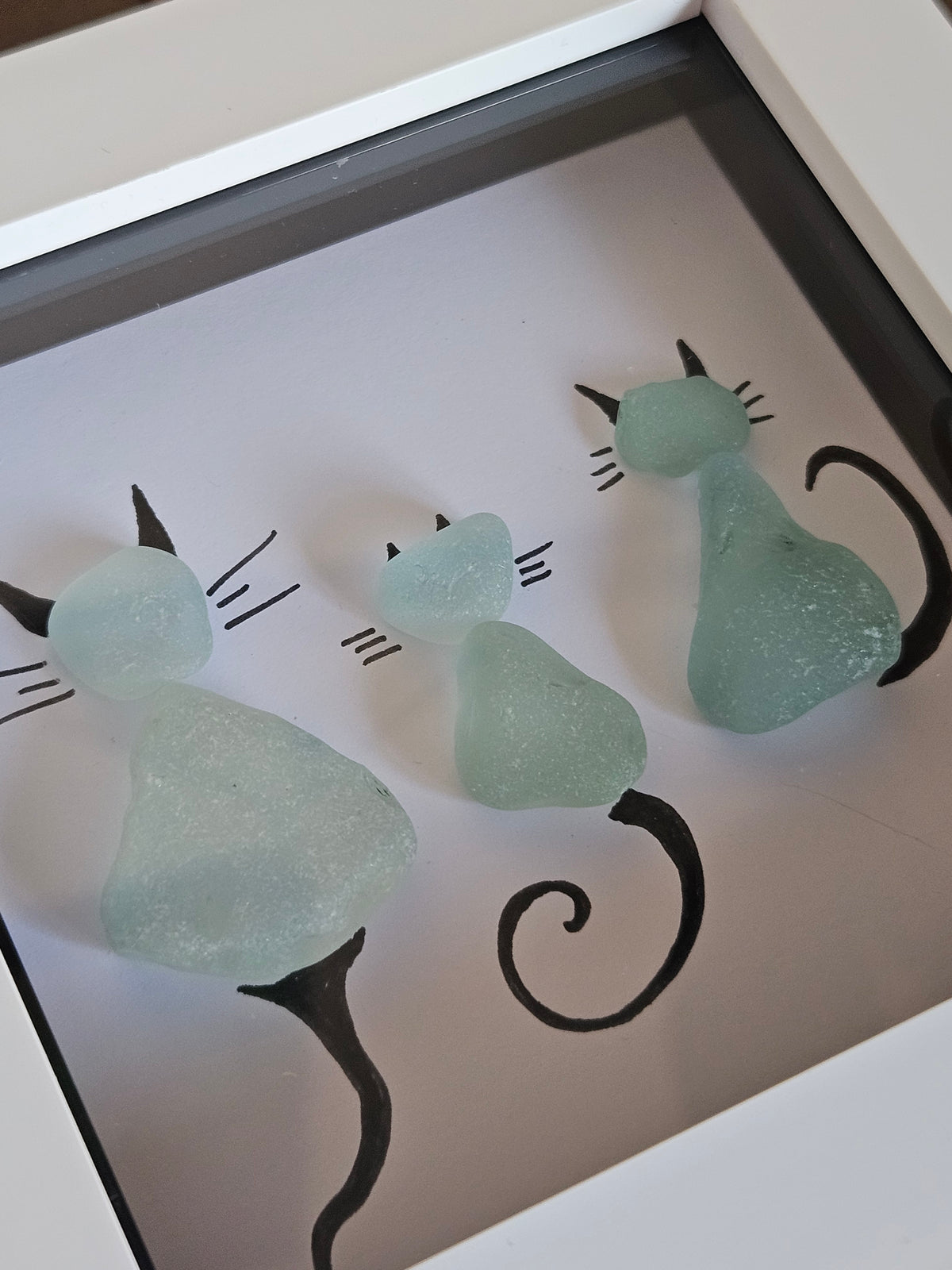 Three sea glass cat figurines in light mint green and pale blue, arranged on a white background inside a framed shadow box with black line art.
