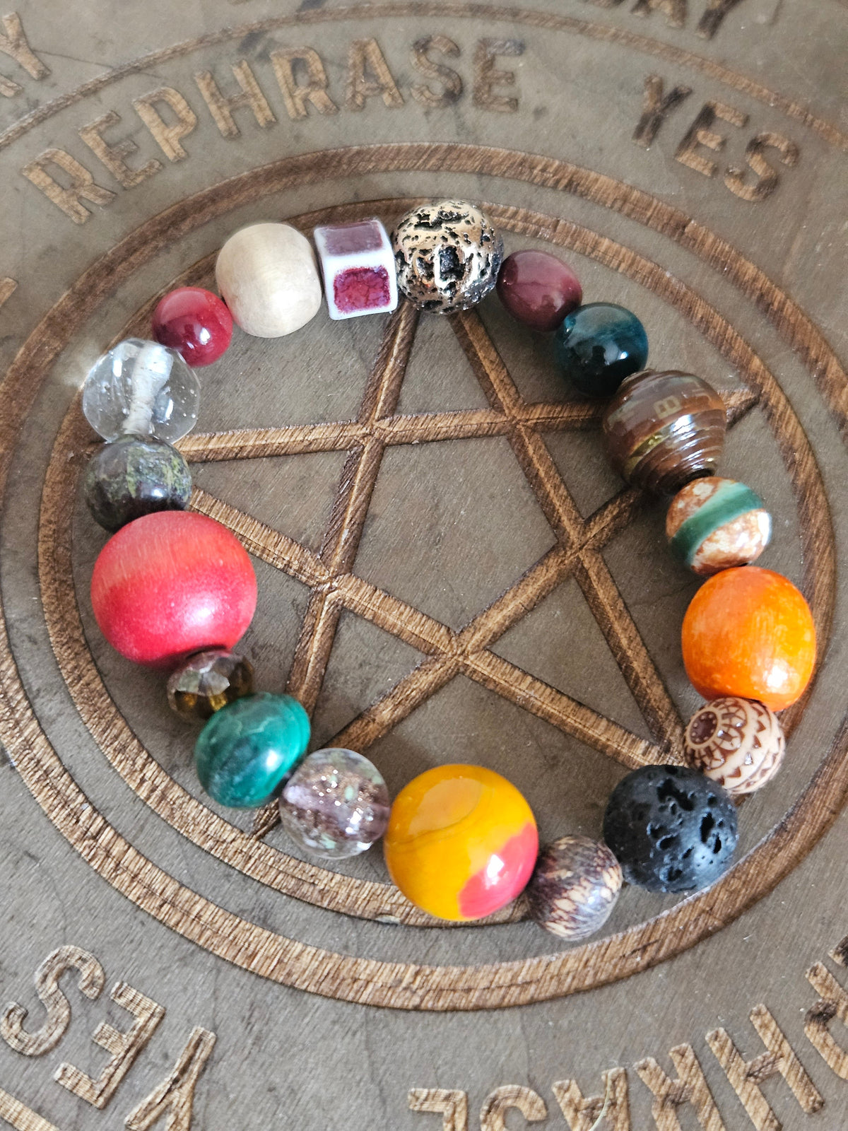 A multicolor beaded bracelet arranged in a circle on a circular wooden board with a pentagram design and engraved words like 'REPHRASE' and 'YES'. The beads include red, orange, yellow, green, black, white, and patterned stones.