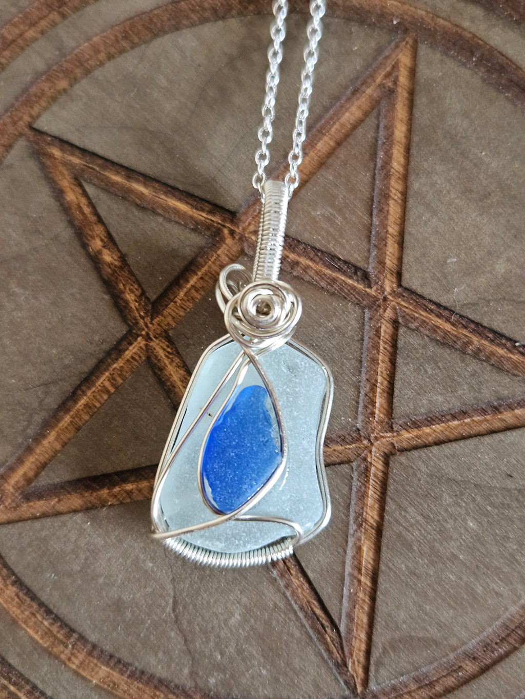 Silver wire wrapped sea glass pendant with blue tones of glass on a wooden pentagram board