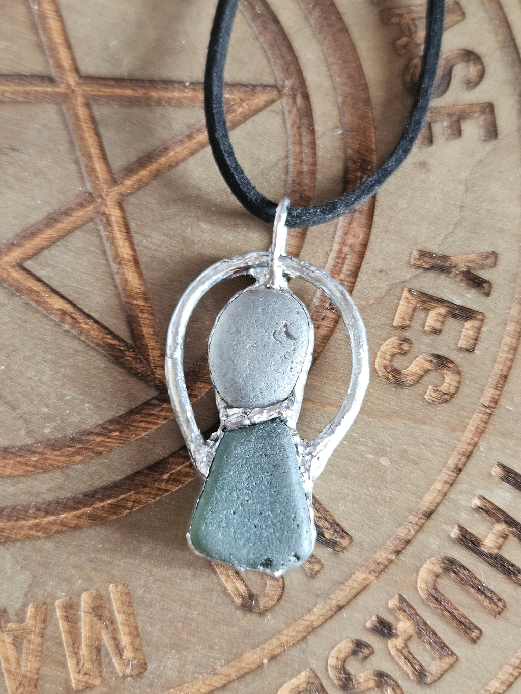 A handmade pendant necklace featuring a silver-toned metal ring with a dual-stone centerpiece, one smooth silver oval and one textured dark gray-green stone, suspended from a black cord. The pendant is displayed on a wooden surface engraved with a pentagram and the word 'THURSDAY'.