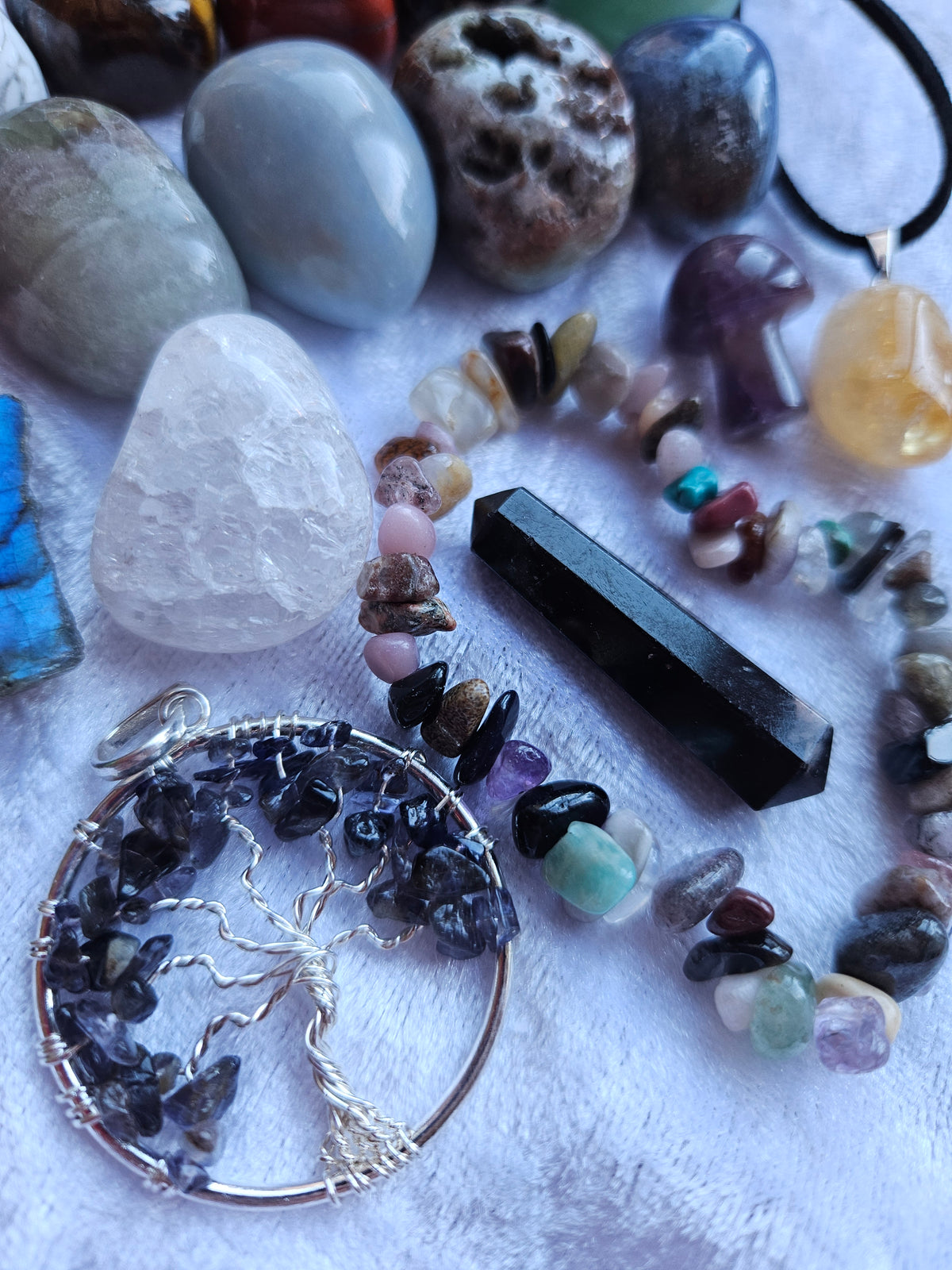A collection of handmade gemstone jewelry and raw stones arranged on a white textured fabric background, featuring a beaded bracelet, a black crystal wand, a silver tree of life pendant filled with dark gemstones, and polished stones including amethyst, rose quartz, and obsidian.