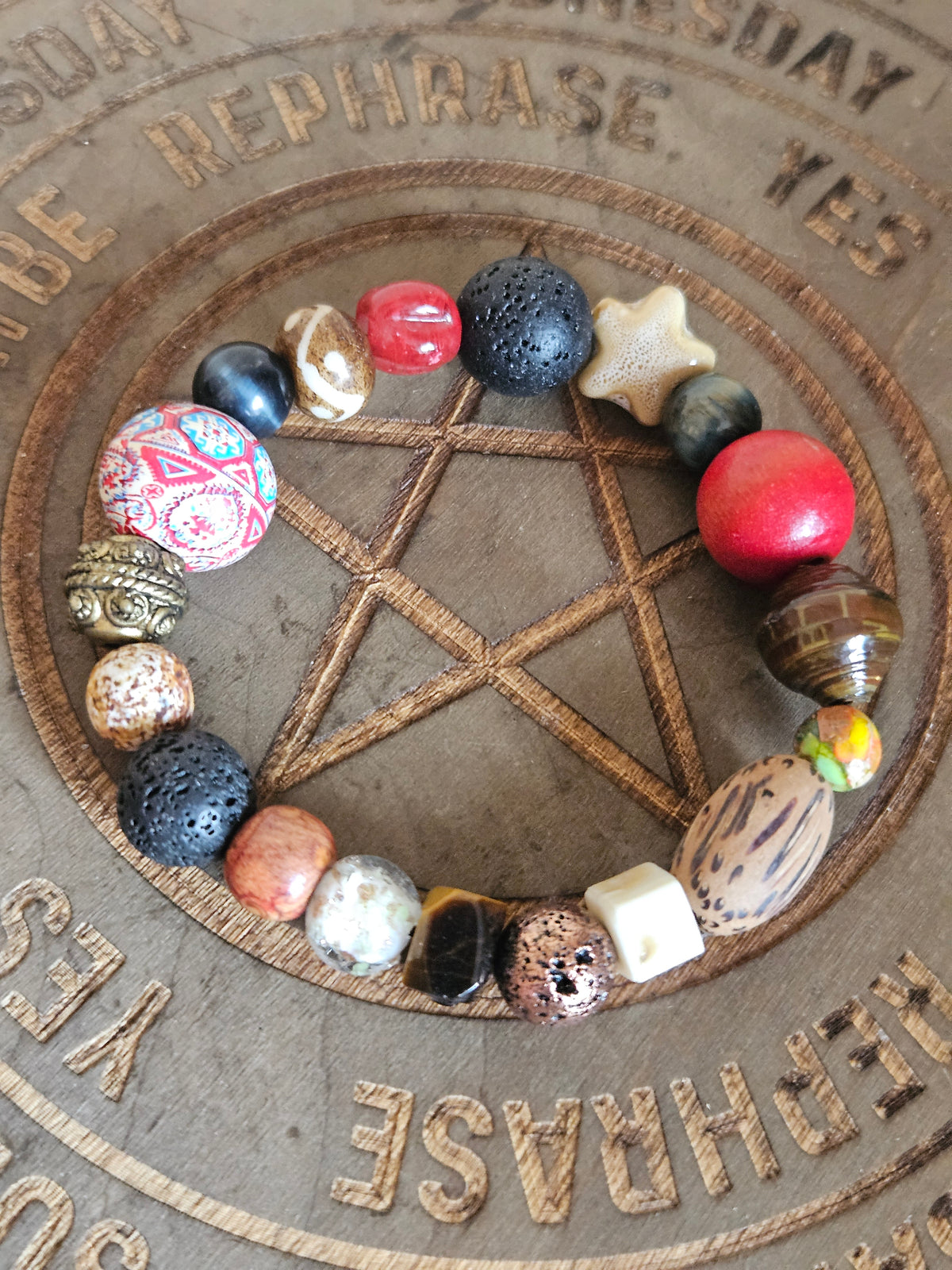 A beaded boho bracelet arranged in a circle on a wooden surface featuring a pentagram design. The bracelet contains a variety of beads including black lava stones, a red round bead, a patterned pink and white bead, a white star bead, and natural wood beads.