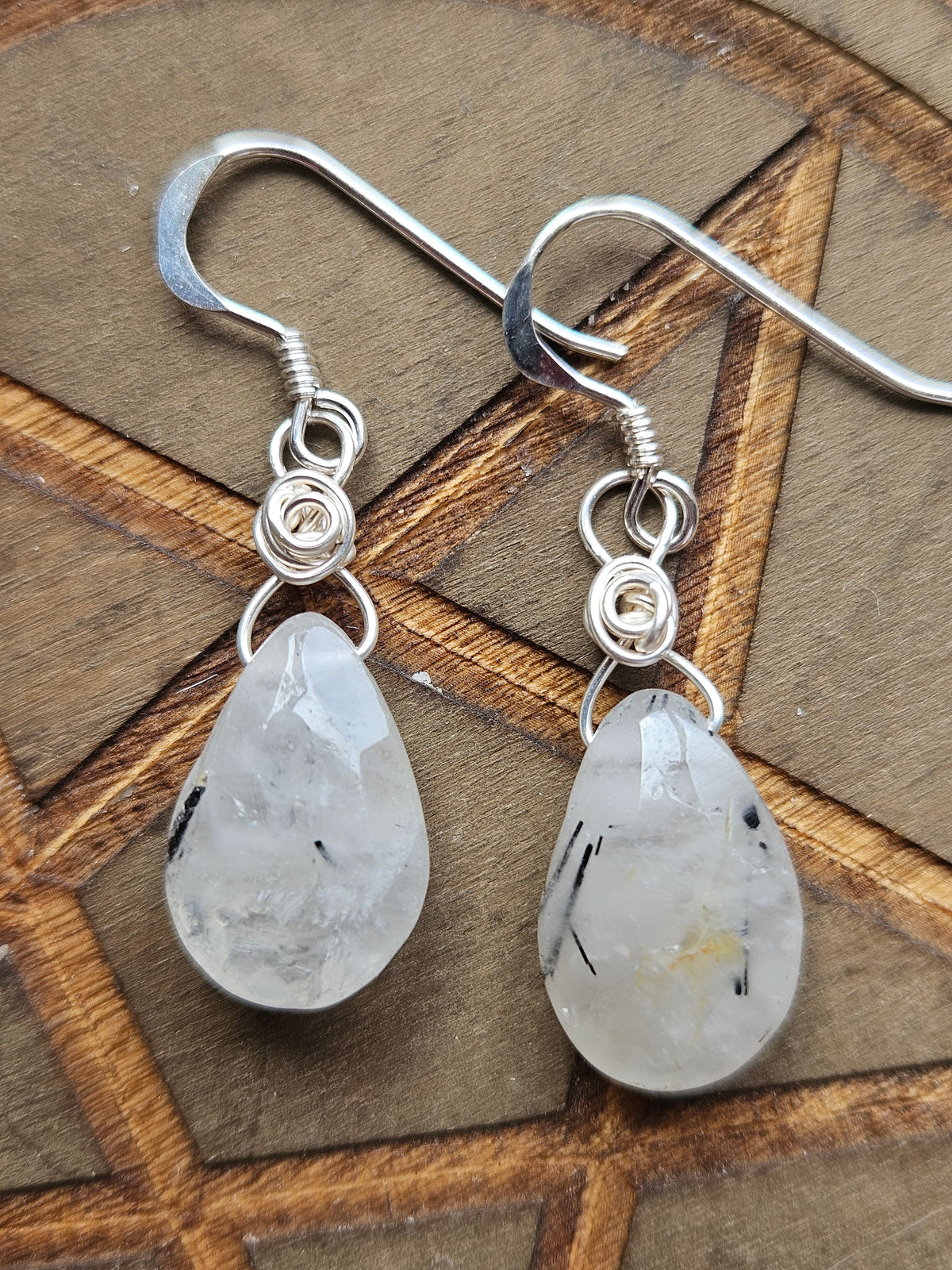 A pair of silver drop earrings with teardrop-shaped Tourmaline In Quartz quartz gemstones, white with black inclusions, displayed on a brown geometric wooden background.