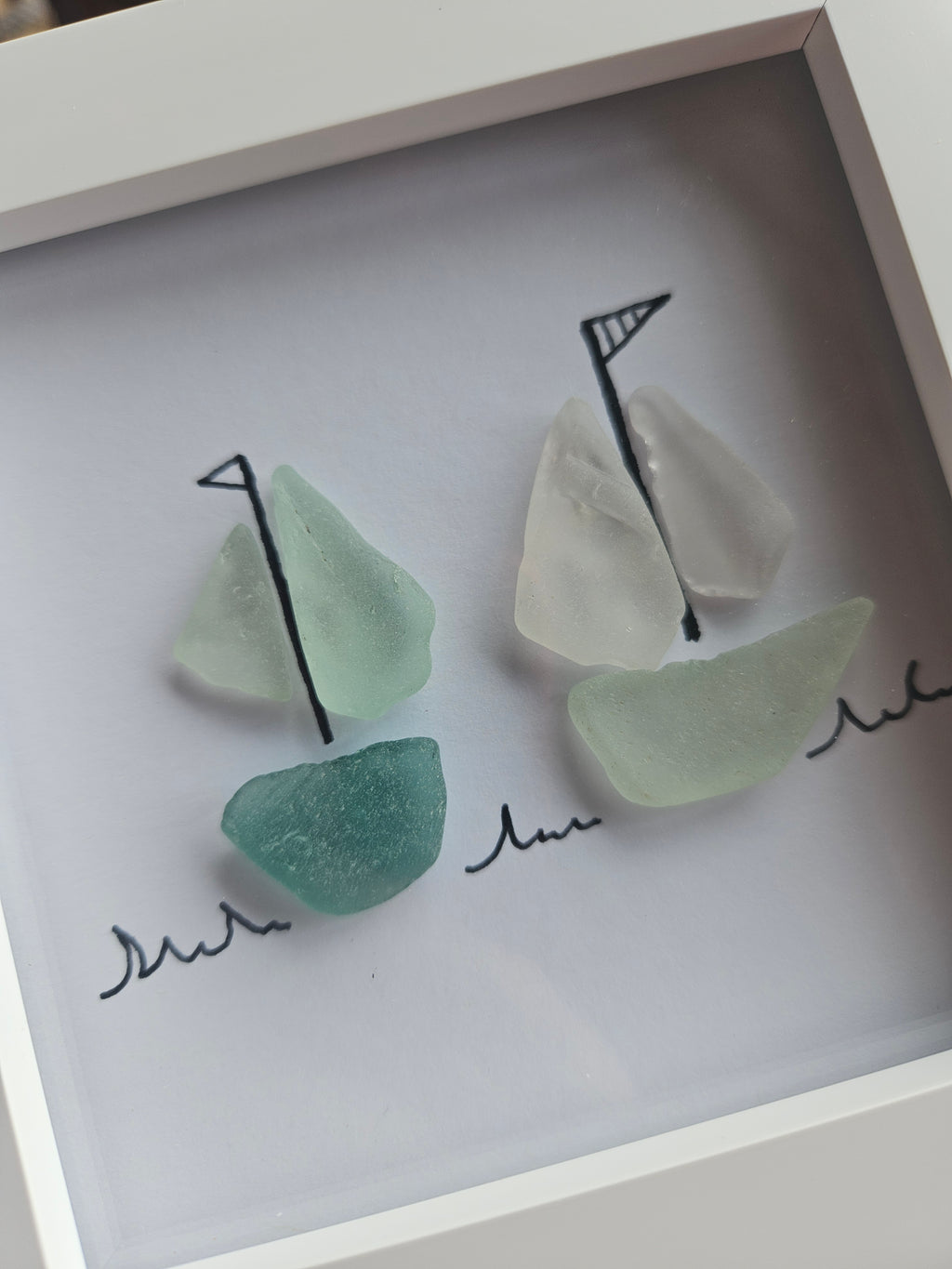 A framed piece of nautical wall art featuring two sea glass sailboats. The boats are constructed from frosted sea glass in shades of mint green, teal, and milky white, with simple black hand-drawn style sails and script wave details on a white background inside a white frame.
