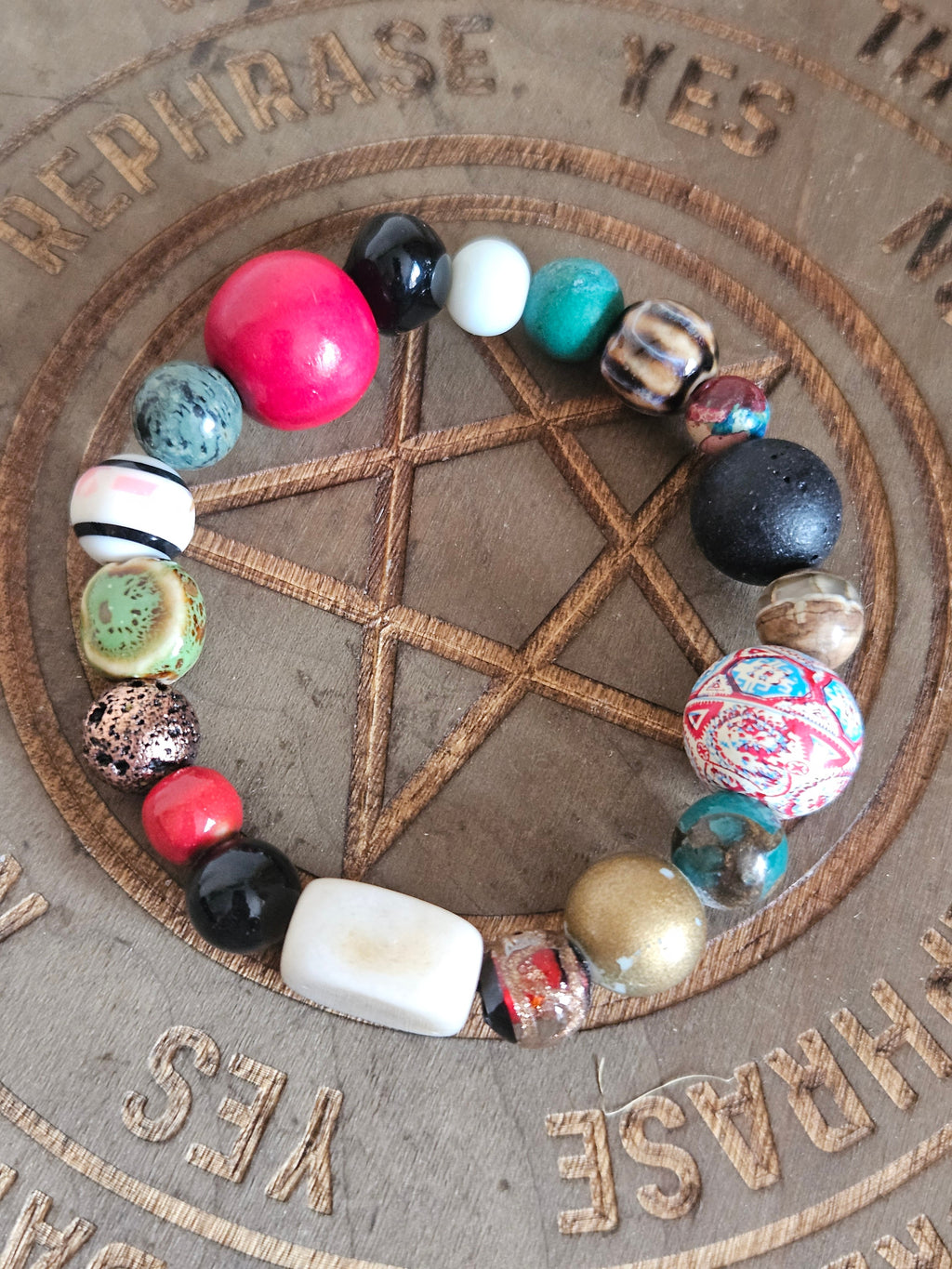 A beaded boho bracelet arranged in a circle on a wooden engraved surface featuring a pentagram and the words 'REPHRASE YES'. The bracelet contains a variety of colorful, patterned, and textured beads including pink, turquoise, black, white, and metallic gold.