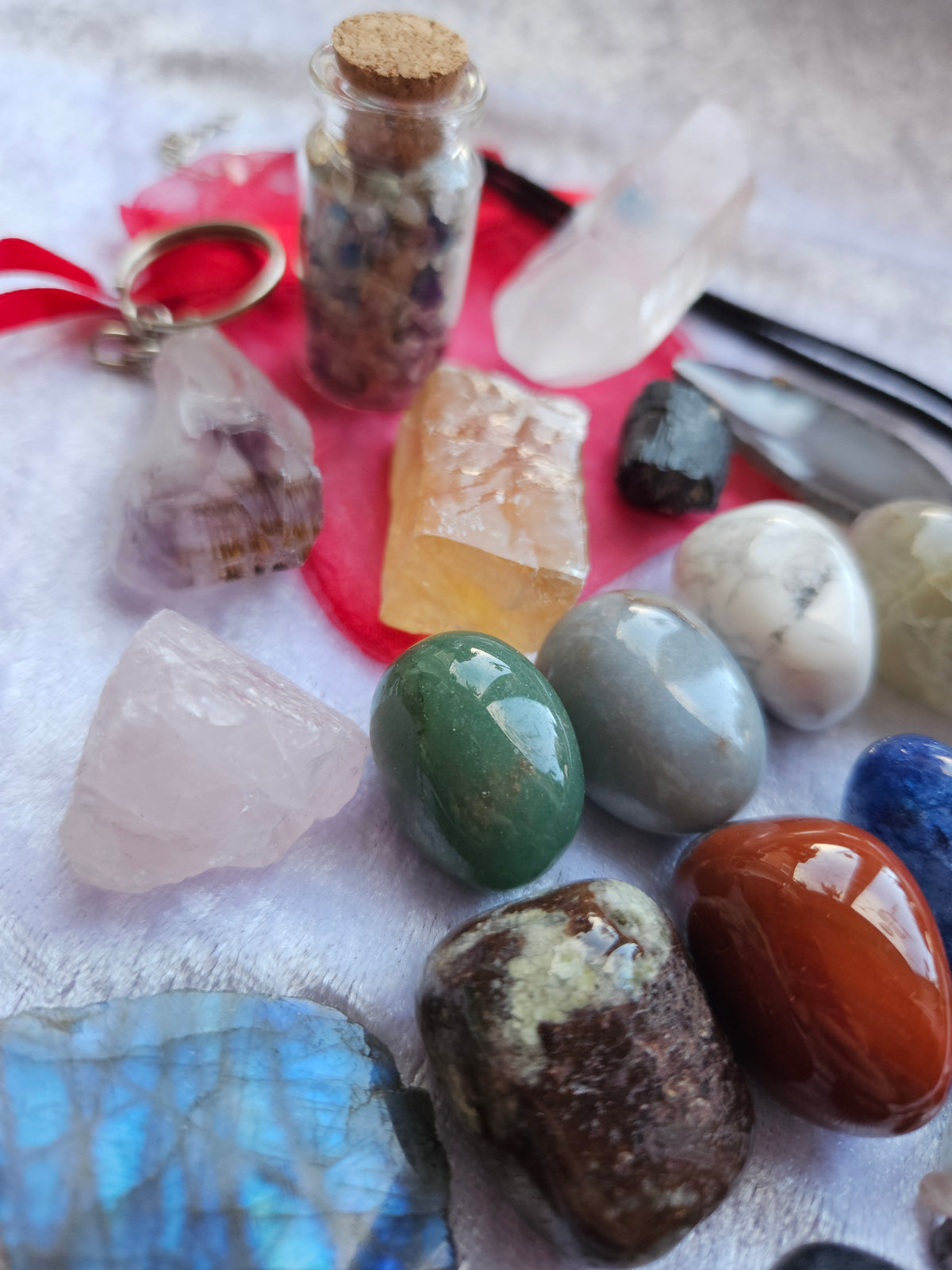 A collection of polished and raw crystals and gemstones arranged on a white textured fabric, including rose quartz, citrine, green jasper, labradorite, and a small glass vial with a cork stopper, alongside a red organza bag and keychain 