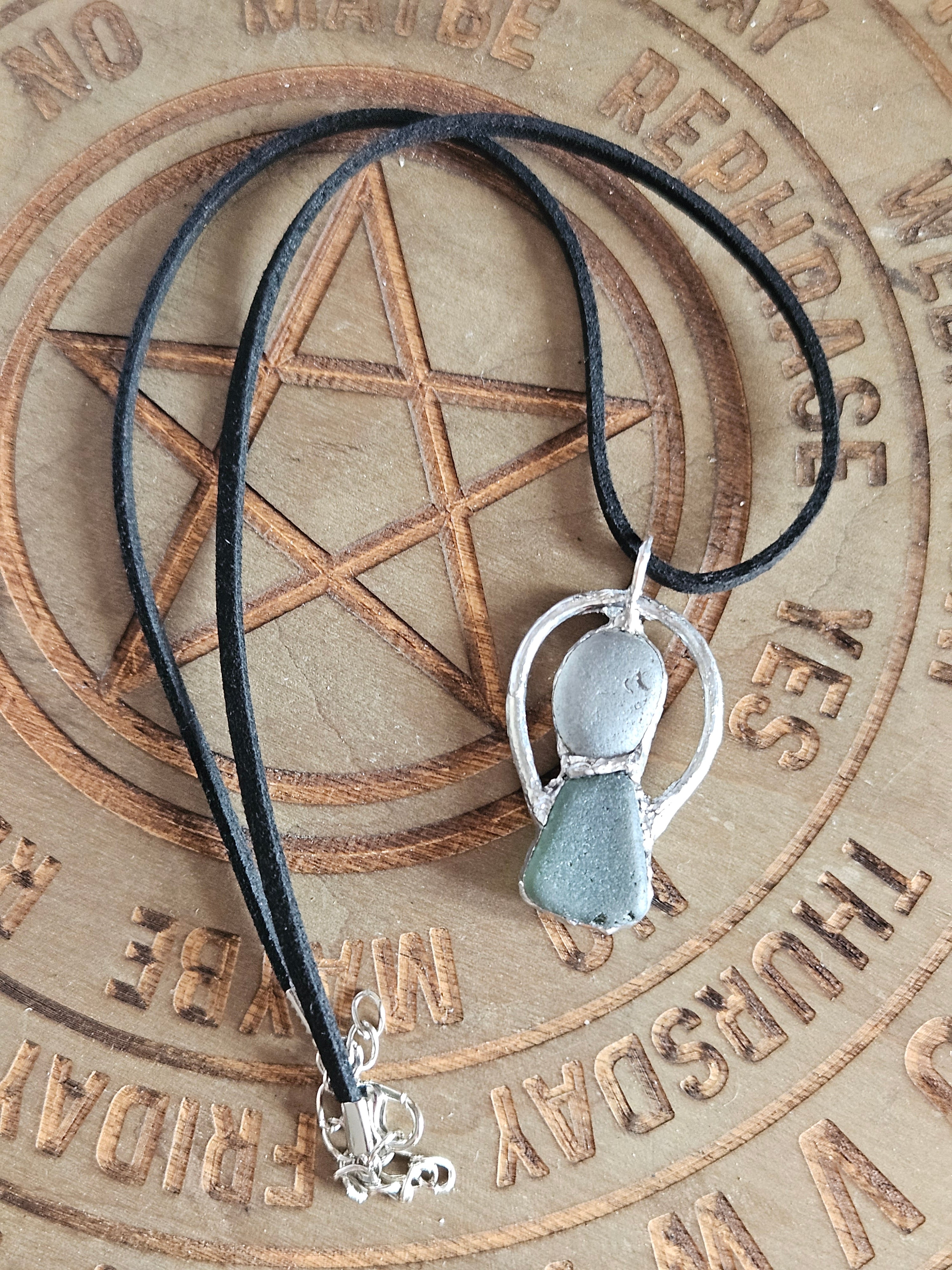 A black cord necklace with a silver-toned pendant featuring two smooth, rounded pale blue-green stones, displayed on a wooden board engraved with a pentagram 