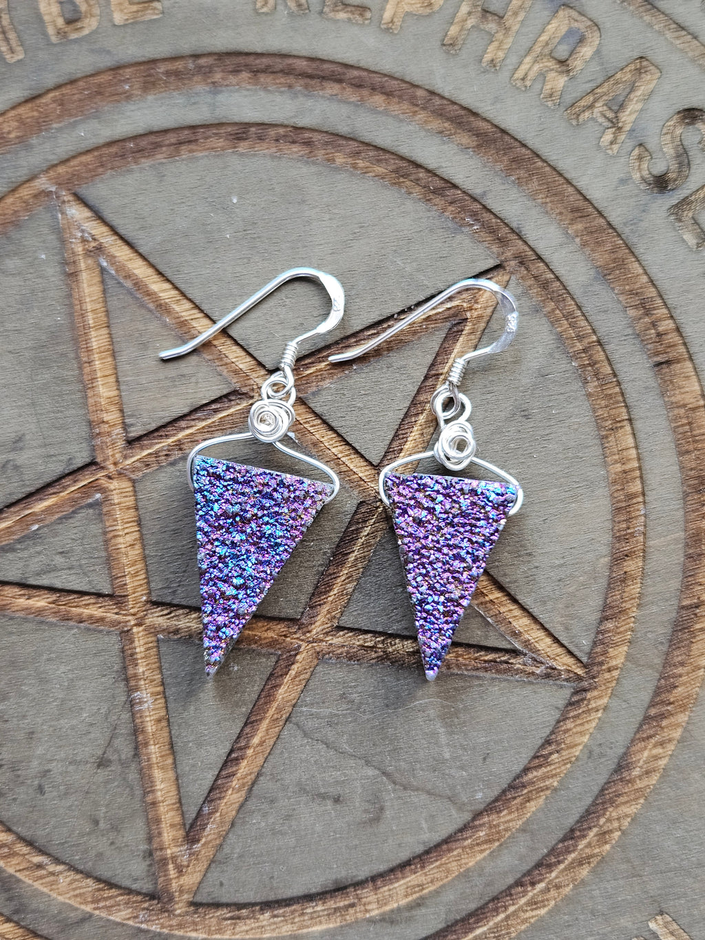 A pair of sterling silver dangle earrings with triangular purple druzy quartz charms, displayed on a wooden pentagram board.