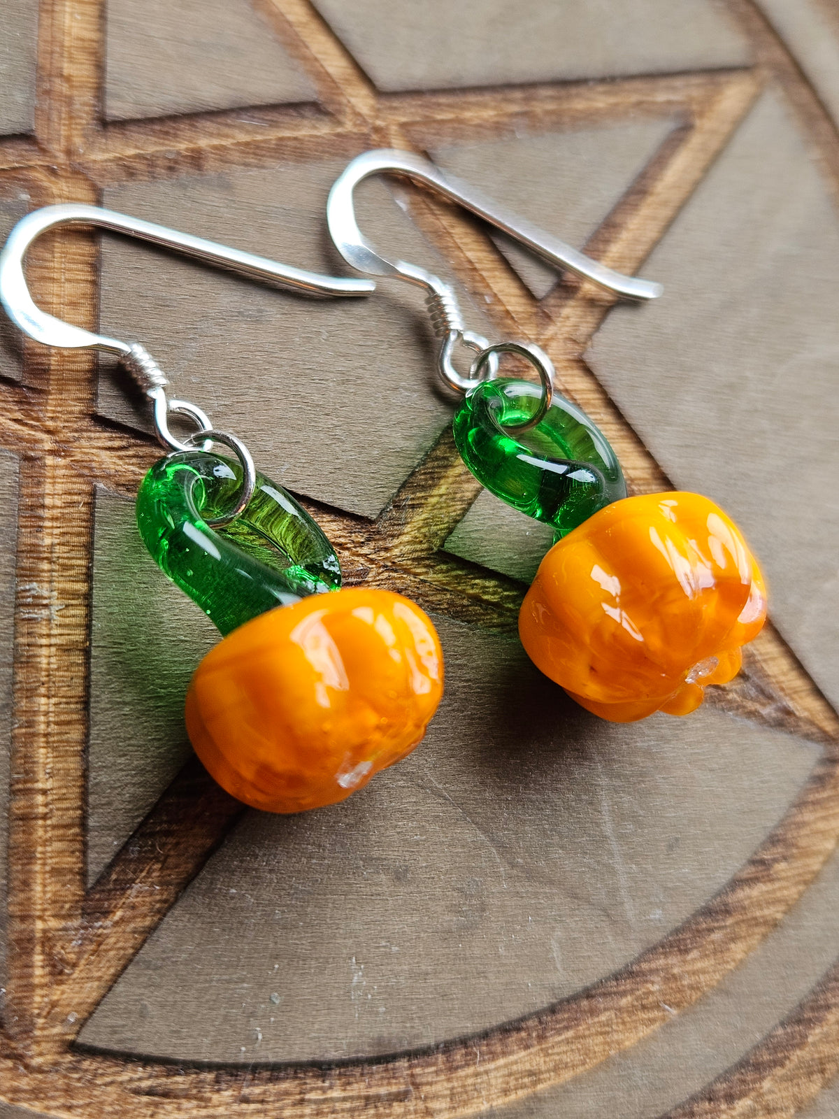 Pumpkin-shaped drop earrings with glossy orange beads and translucent green beads, suspended from silver fishhook wires, displayed on a wooden surface.