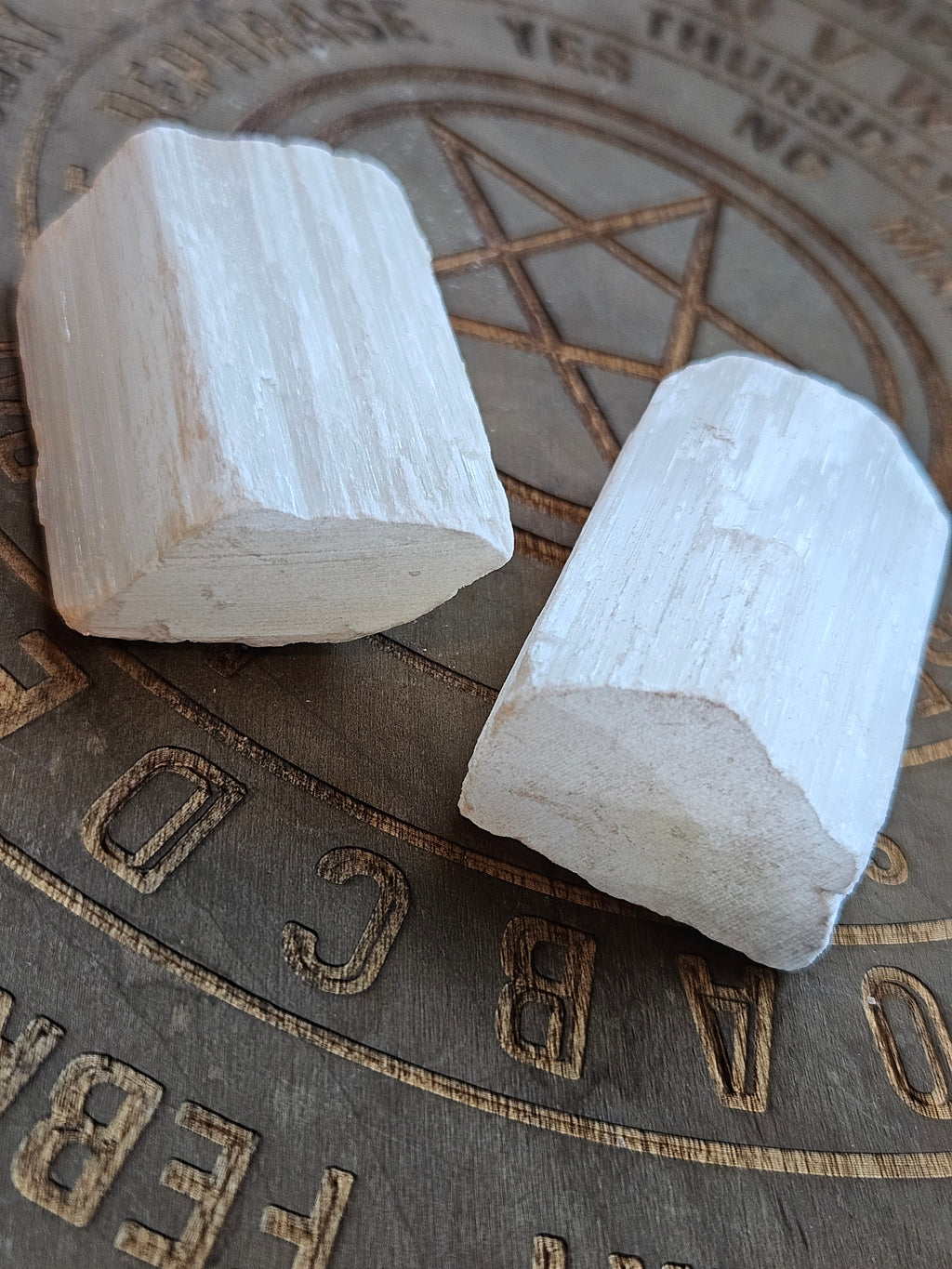Two white rectangular Selenite chunks with a rough, textured surface resting on a dark metallic surface with engraved symbols and letters.