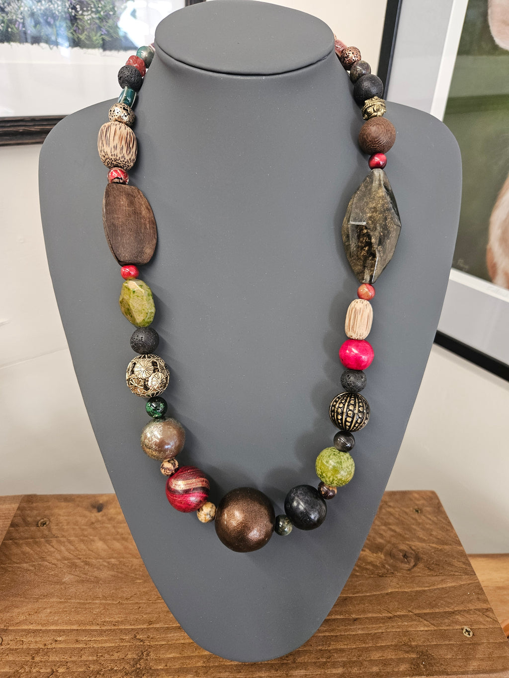 A chunky beaded boho necklace made of mixed natural materials, including wood, stone, and colorful beads, displayed on a matte gray jewelry bust.