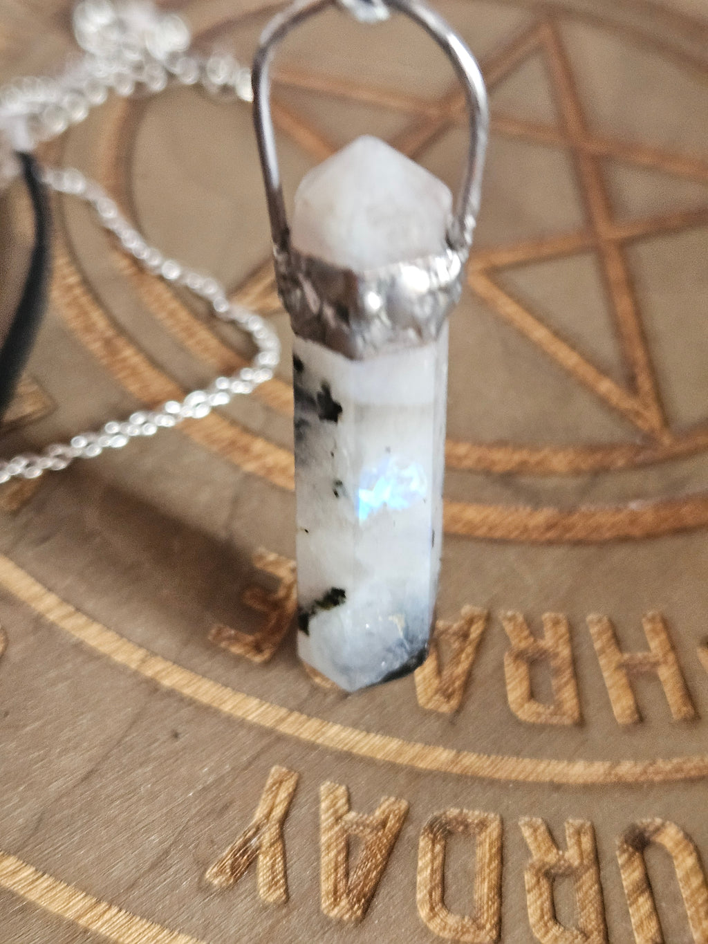 A polished rainbow moonstone crystal pendant with a silver metal loop and chain, resting on a wooden surface with a pentagram design.