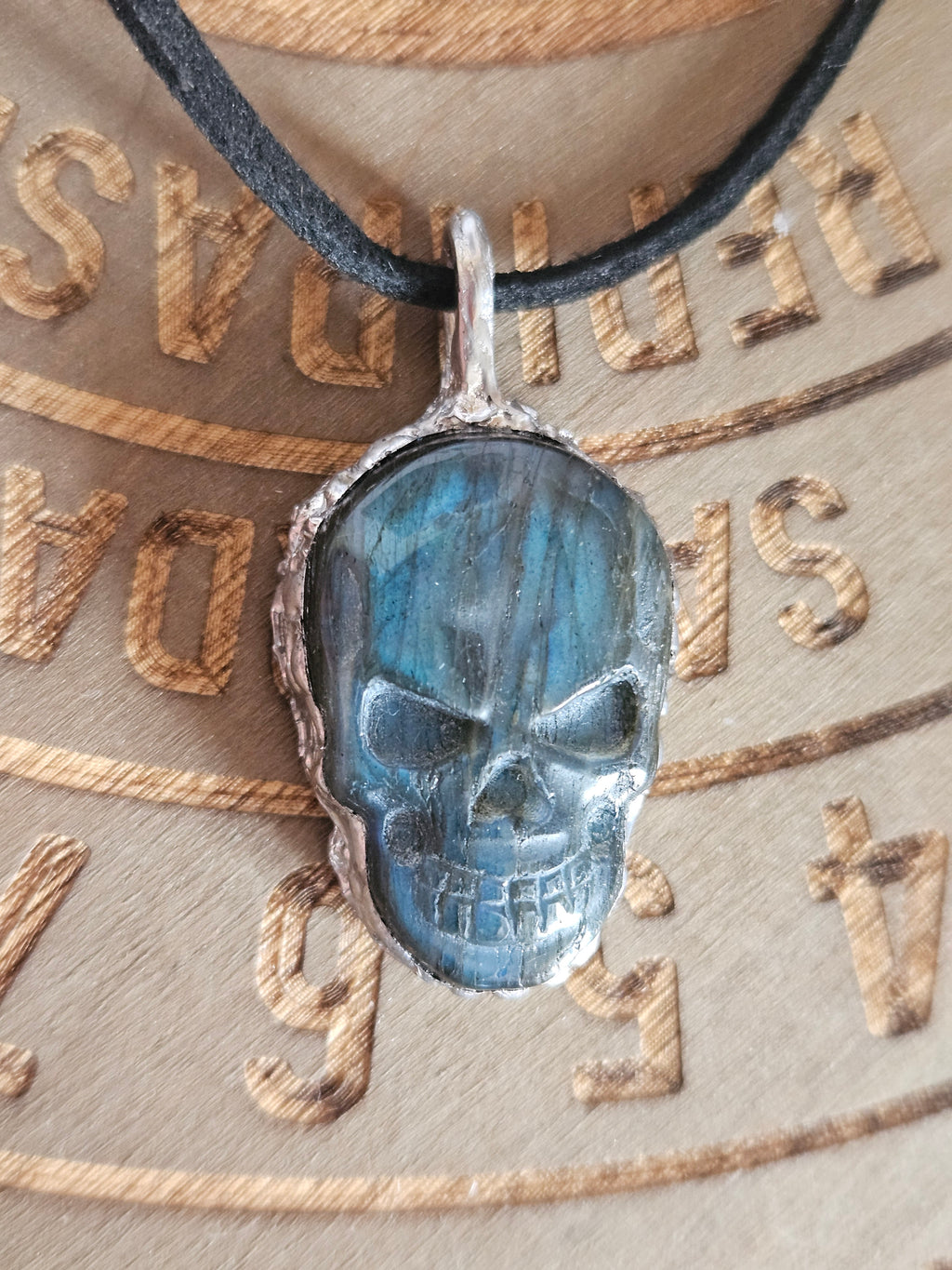 A skull-shaped pendant with a dark blue-gray stone face and a silver-toned textured border, hanging from a black cord over a beige printed background with engraved letters.