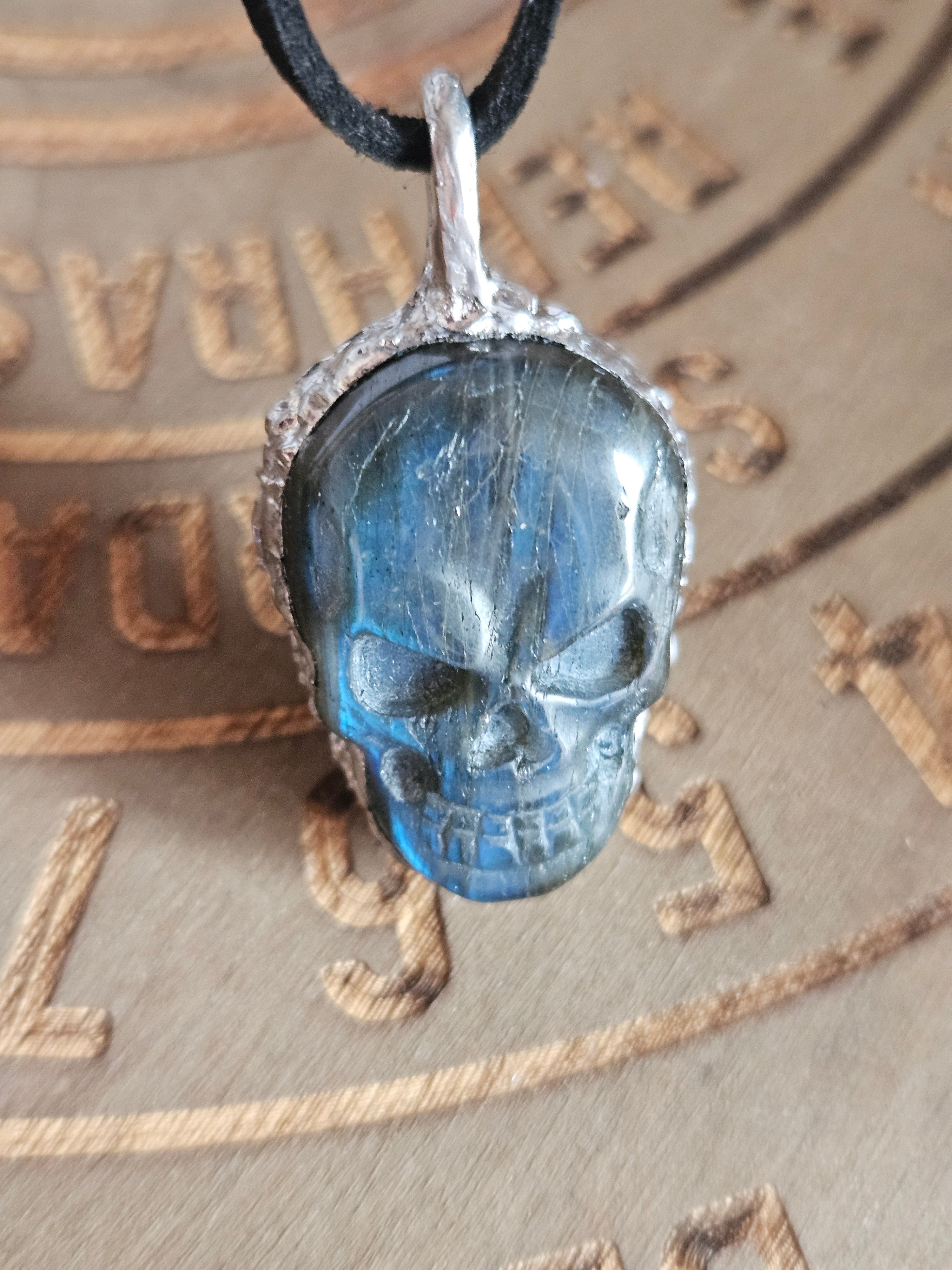 A carved blue labradorite skull pendant with a silver-toned setting and a black cord, shown close-up on a textured brown background.