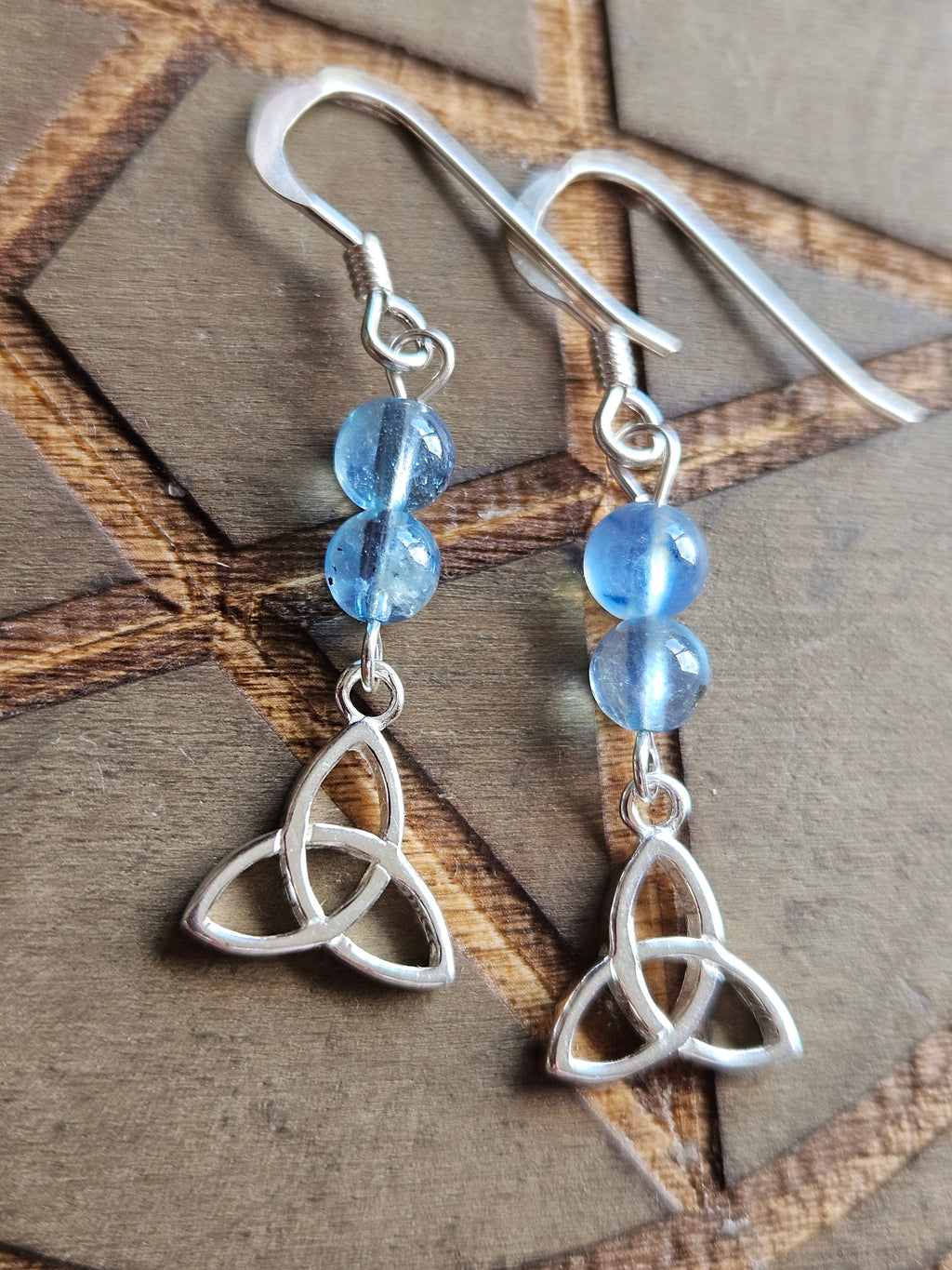 A pair of sterling silver dangle earrings with two light blue Aquamarine beads and a triquetra knot charm, displayed on a textured brown background.