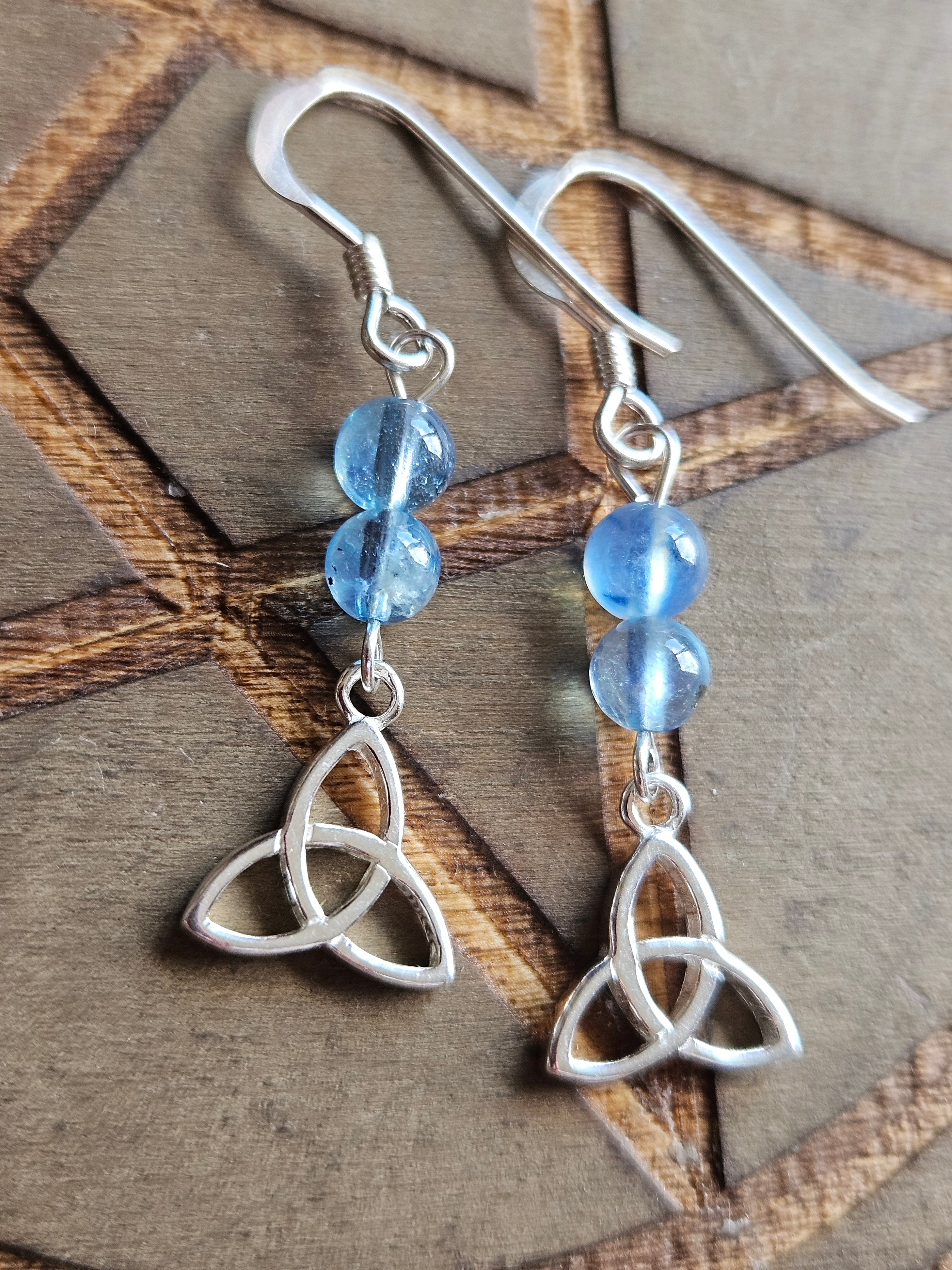 A pair of sterling silver dangle earrings with two light blue Aquamarine beads and a triquetra knot charm, displayed on a textured brown background.