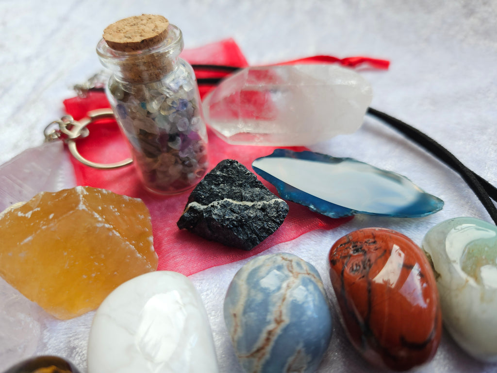A crystal and gemstone set arranged on a white textured surface, including polished stones, a raw black stone, a blue-green agate slice, a small glass vial with a cork stopper and key ring, and a red cloth.