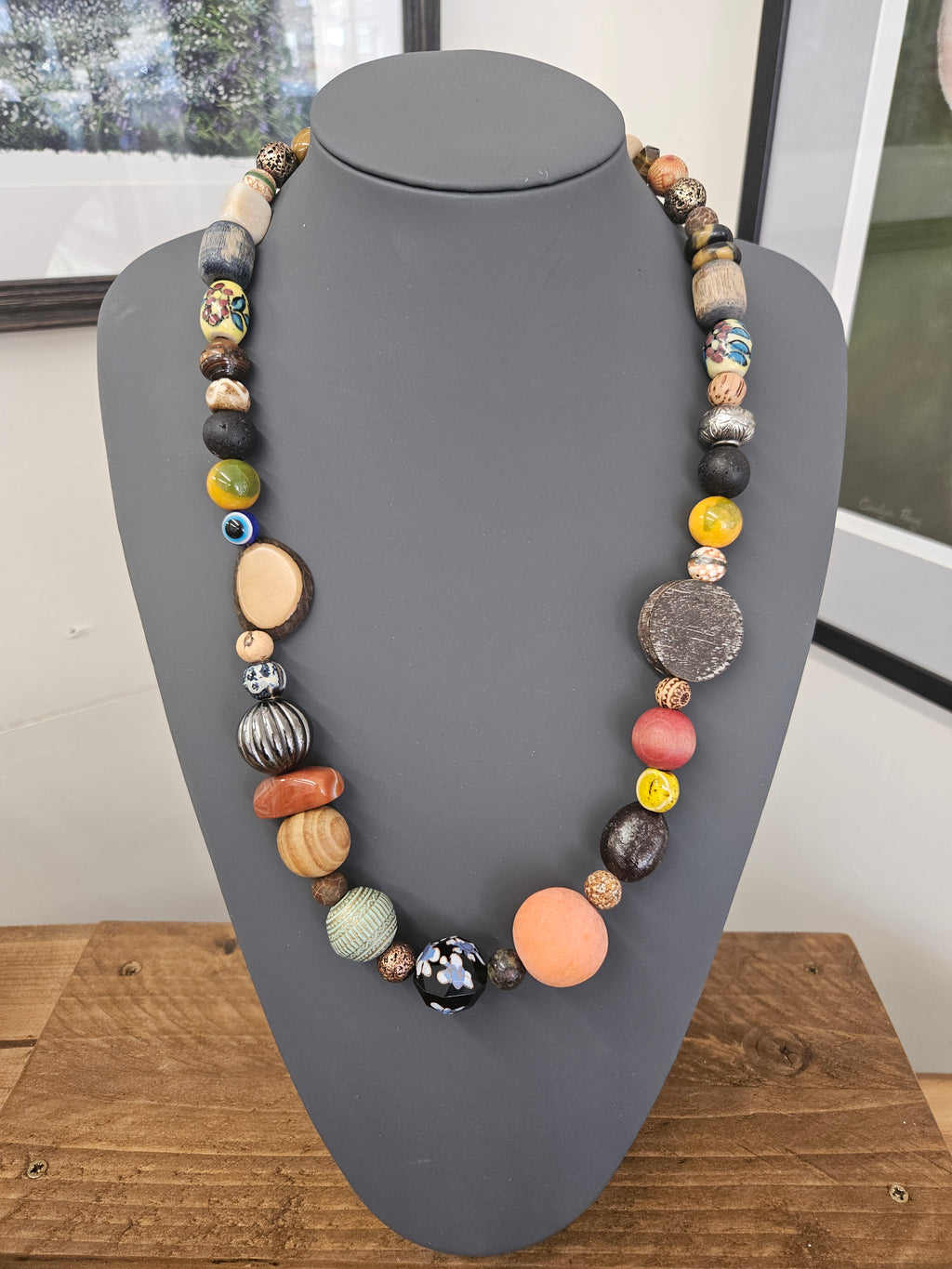 A handmade beaded boho necklace displayed on a matte gray jewelry bust, featuring a variety of colorful and textured beads including wood, stone, glass, and metallic elements.