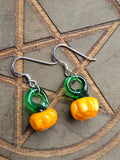 A pair of handmade dangle earrings featuring glossy green and orange glass pumpkin beads on silver fishhook wires, displayed on a dark wood background with an engraved pentagram.
