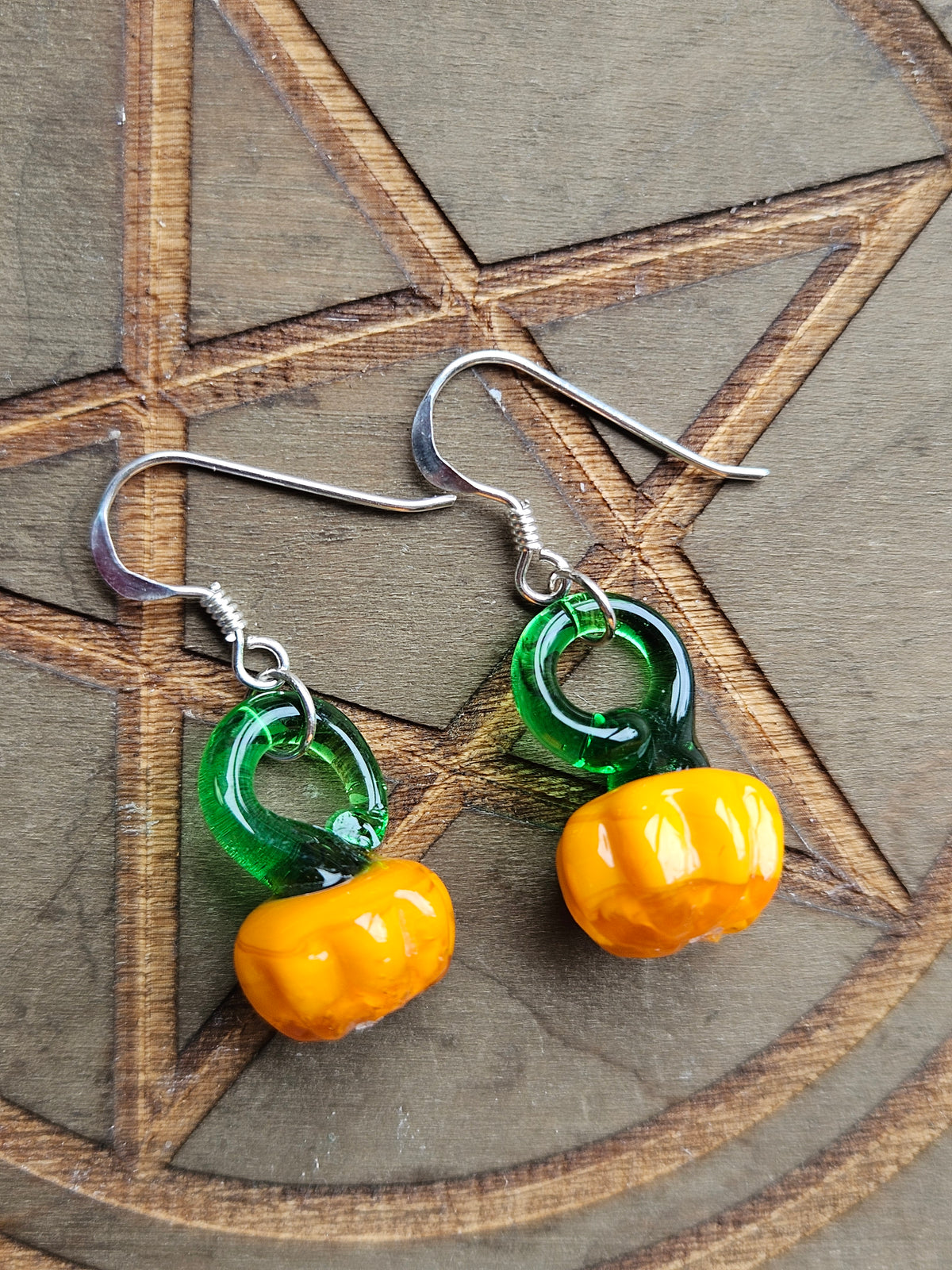 A pair of handmade dangle earrings featuring glossy green and orange glass pumpkin beads on silver fishhook wires, displayed on a dark wood background with an engraved pentagram.