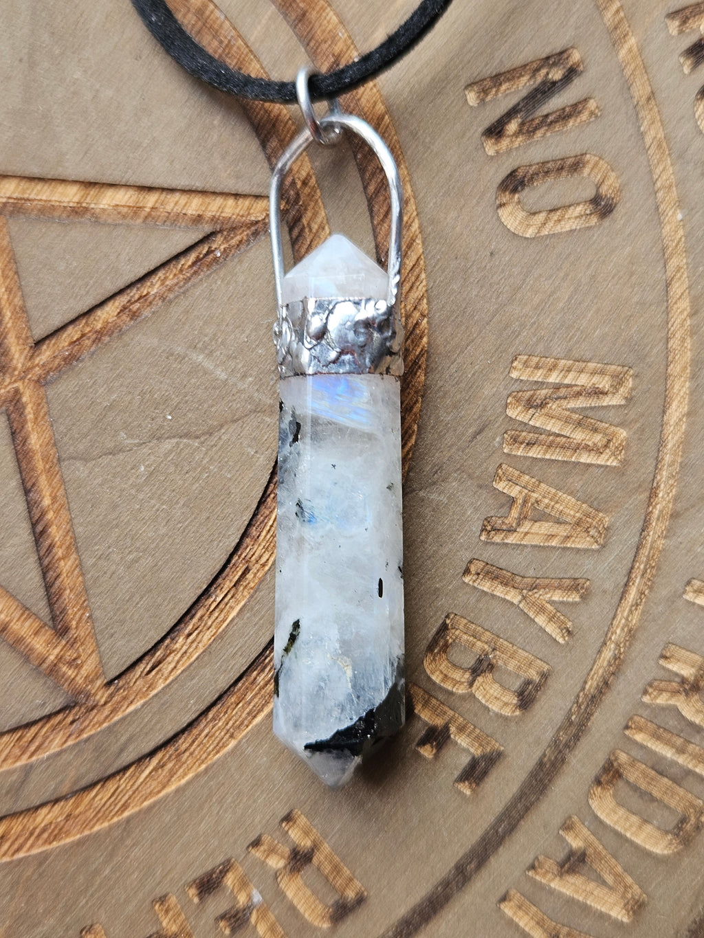 A polished rainbow moonstone point pendant with a silver metal cap and loop, resting on a brown engraved wooden surface.