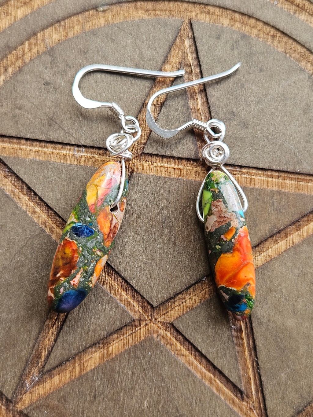 A pair of handmade dangle earrings with silver fishhook wires and colorful marbled Sediment Jasper beads in orange, blue, green, and yellow, displayed on a wooden pentagram board.