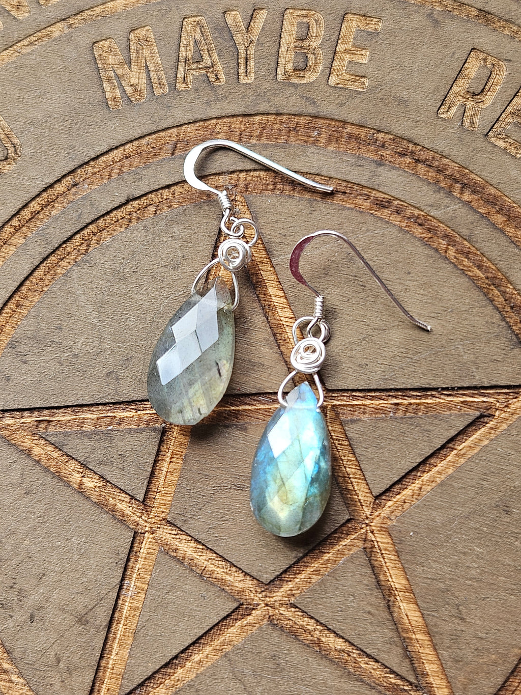 A pair of teardrop-shaped labradorite dangle earrings with silver wire-wrapping and sterling silver fishhook backs, displayed on a wooden pentagram board.
