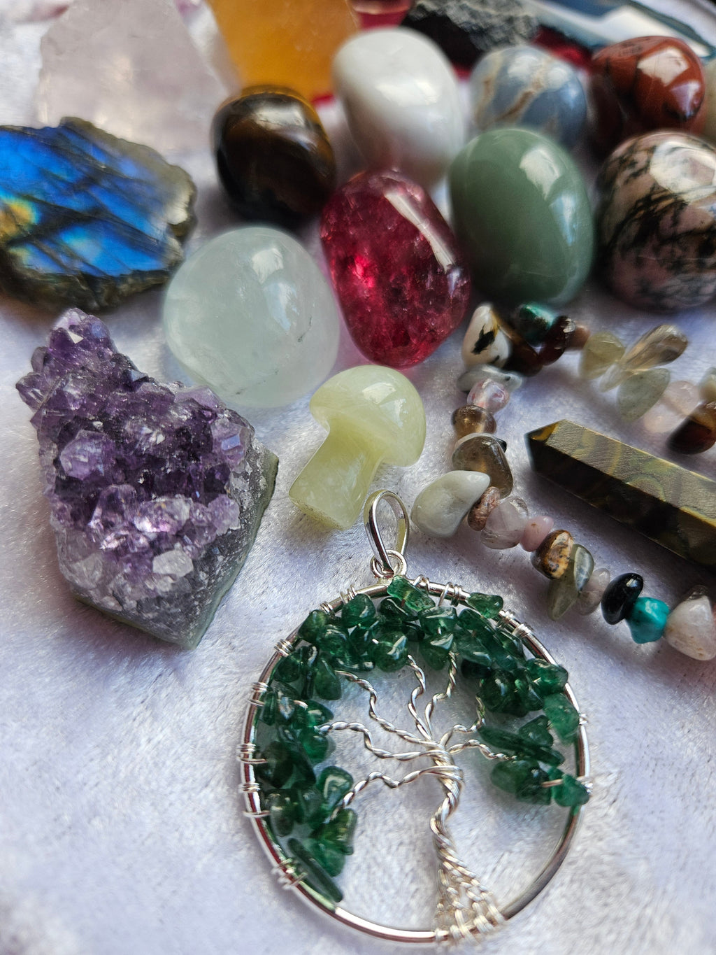 A collection of crystal jewelry and raw gemstones arranged on a white textured fabric background, featuring a silver tree-of-life pendant made of green crystals, polished stones, and raw crystals.