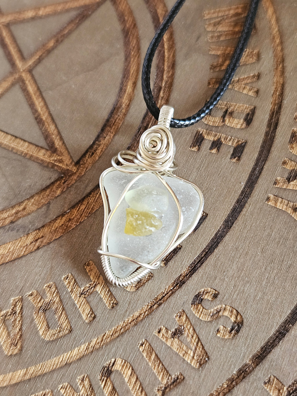 A close-up of a silver wire-wrapped pendant necklace with a translucent white sea glass and a small yellow inlay, hanging from a black cord over a engraved wooden surface.