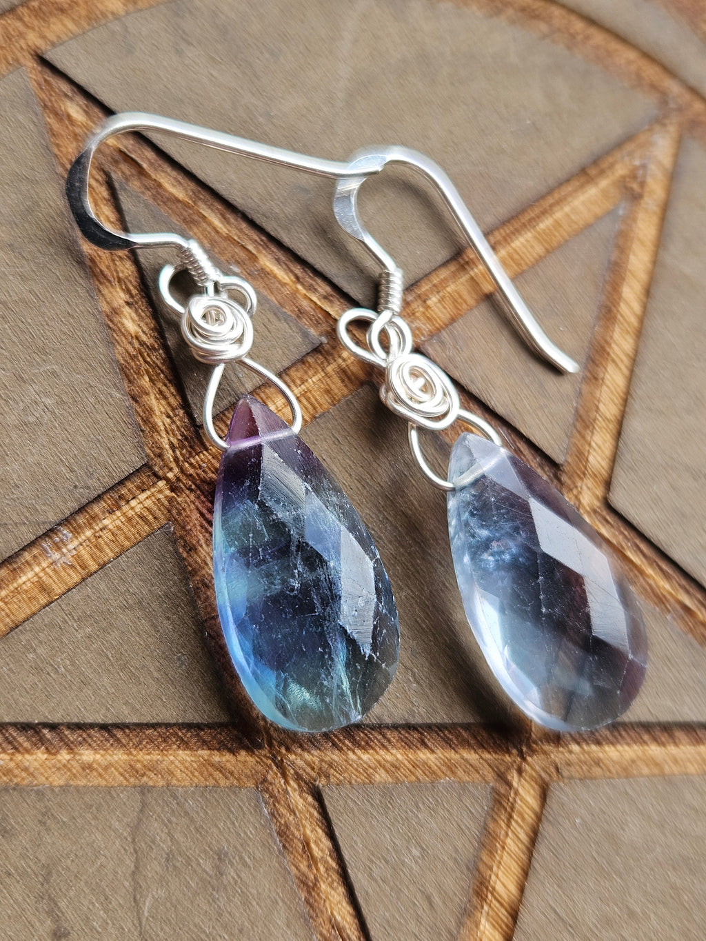 A pair of handmade silver wire-wrapped drop earrings with faceted teardrop-shaped blue and purple Fluorite gemstone pendants, displayed on a brown wooden background with a pentagram design.