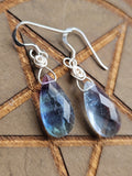 A pair of handmade silver wire-wrapped drop earrings with faceted teardrop-shaped blue and purple Fluorite gemstone pendants, displayed on a brown wooden background with a pentagram design.