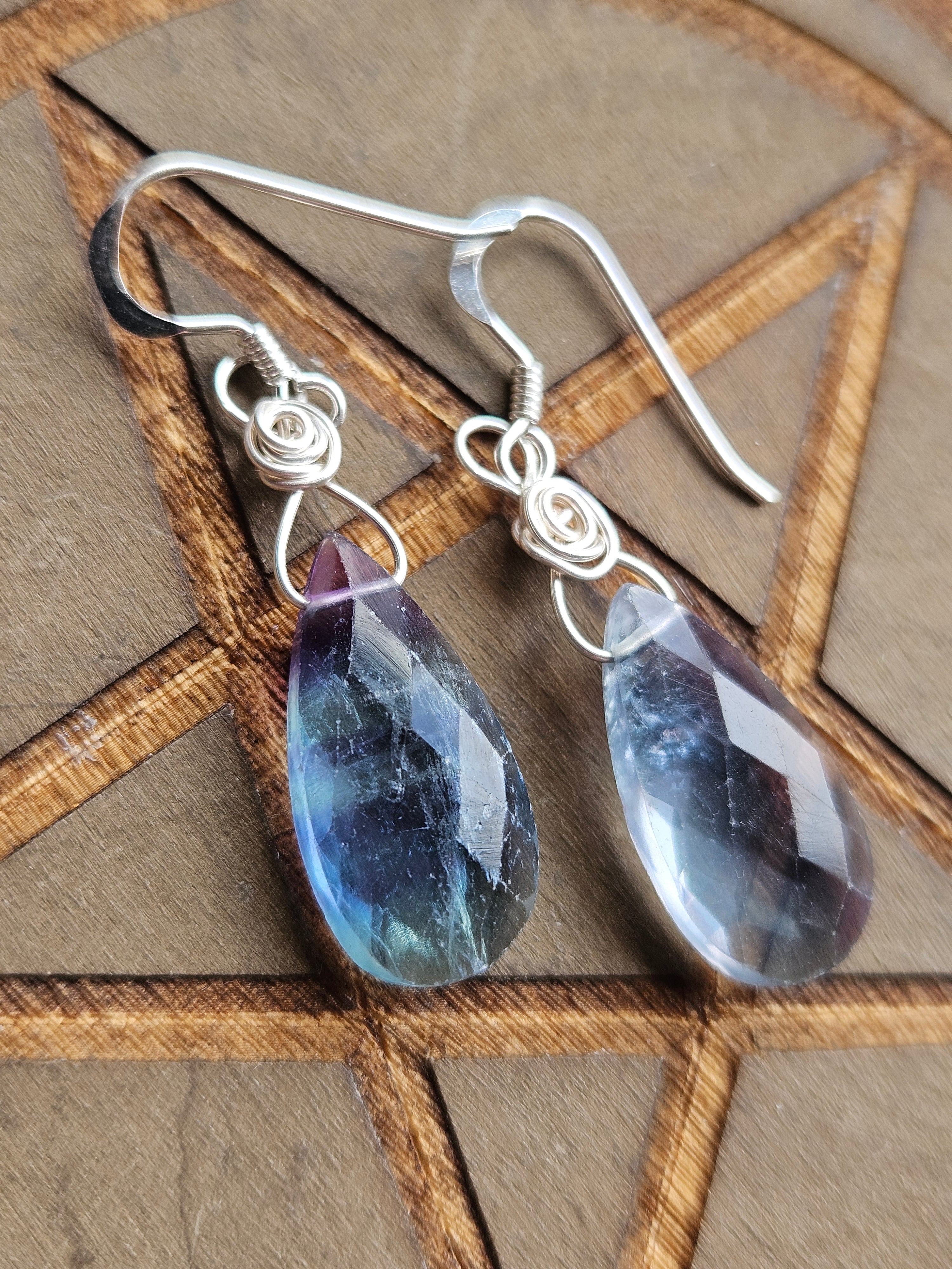 A pair of handmade silver wire-wrapped drop earrings with faceted teardrop-shaped blue and purple Fluorite gemstone pendants, displayed on a brown wooden background with a pentagram design.