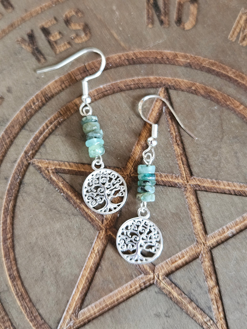 A pair of bohemian-style dangle earrings featuring sterling silver tree of life charms and small green Emerald stones, displayed on a wooden pentagram background.