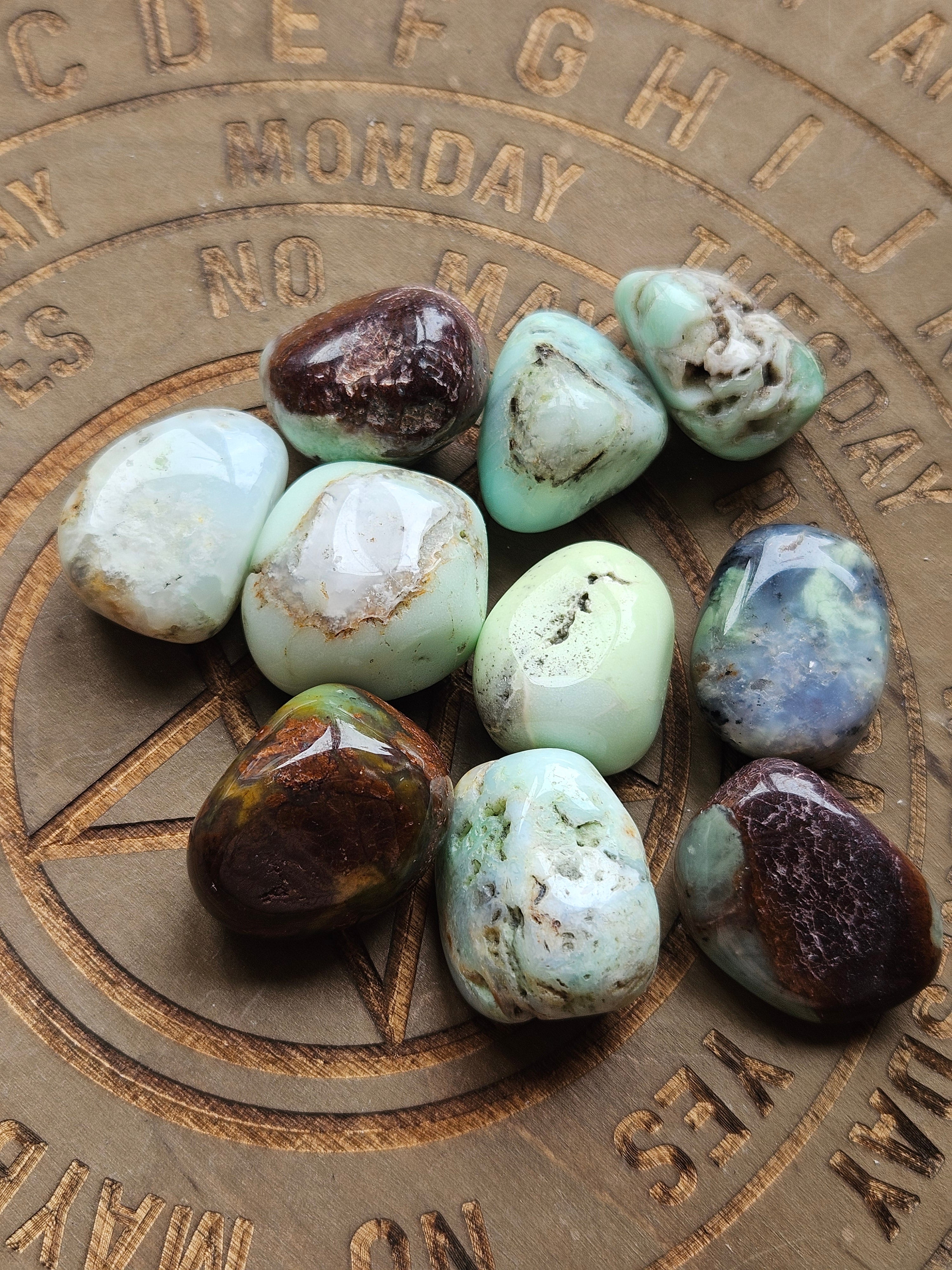 Nine polished tumbled gemstones in various earthy colors and patterns, arranged on a circular brown wooden board engraved with days of the week and a pentagram.