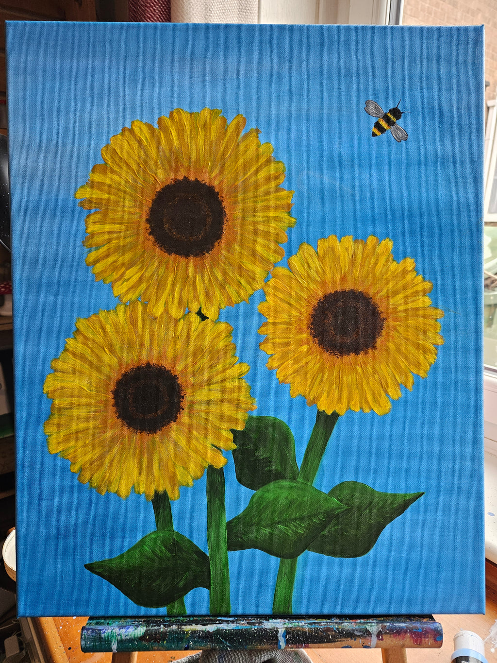 Three sunflowers with a bee on a blue background displayed on easel 