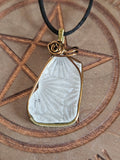 A white textured sea glass pendant wrapped in bronze-toned wire, hanging from a black cord, displayed on a wooden surface with a pentagram design.
