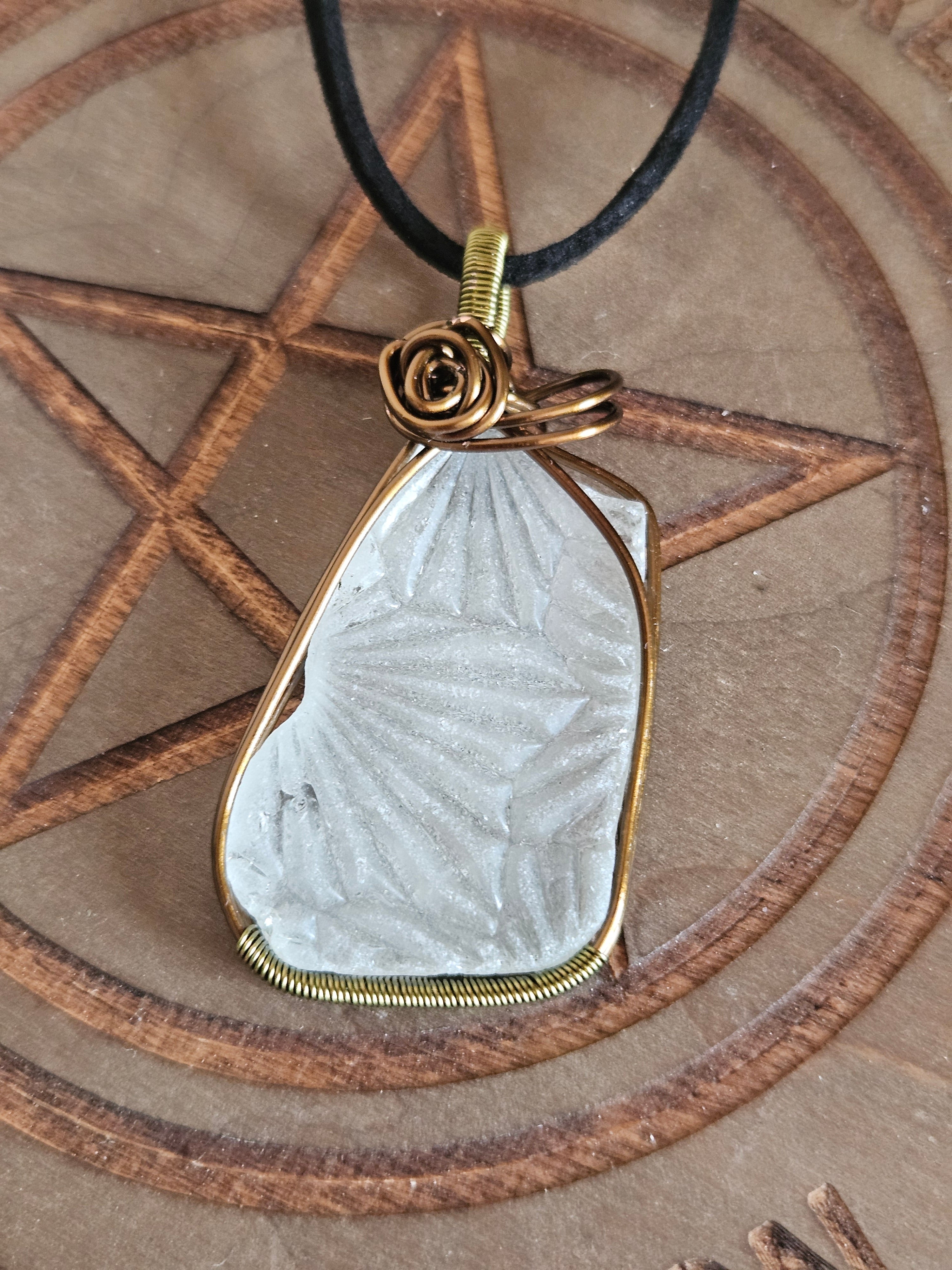 A white textured sea glass pendant wrapped in bronze-toned wire, hanging from a black cord, displayed on a wooden surface with a pentagram design.