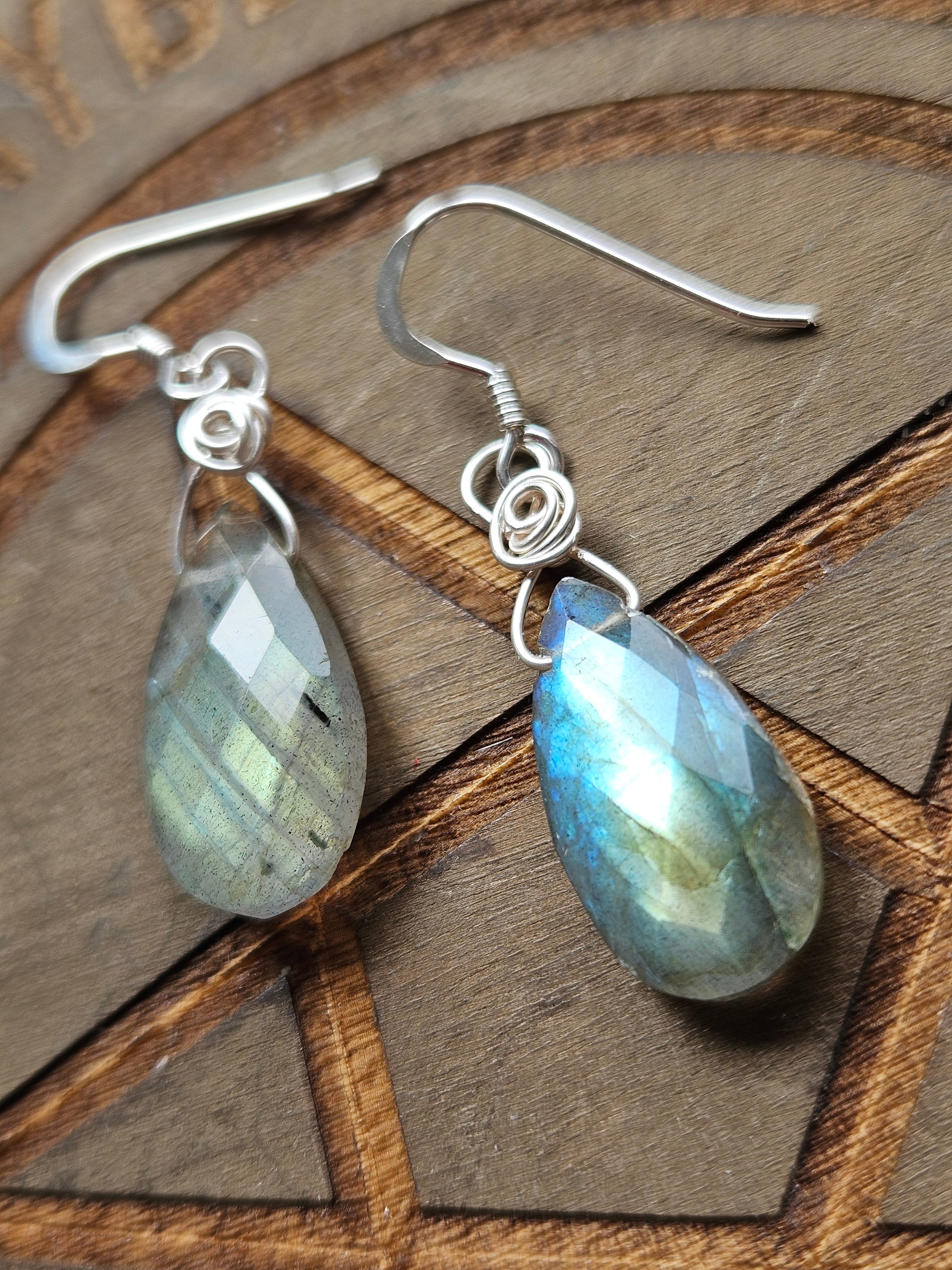 A pair of teardrop-shaped labradorite dangle earrings with silver wire-wrapped hooks, displayed on a wooden background.