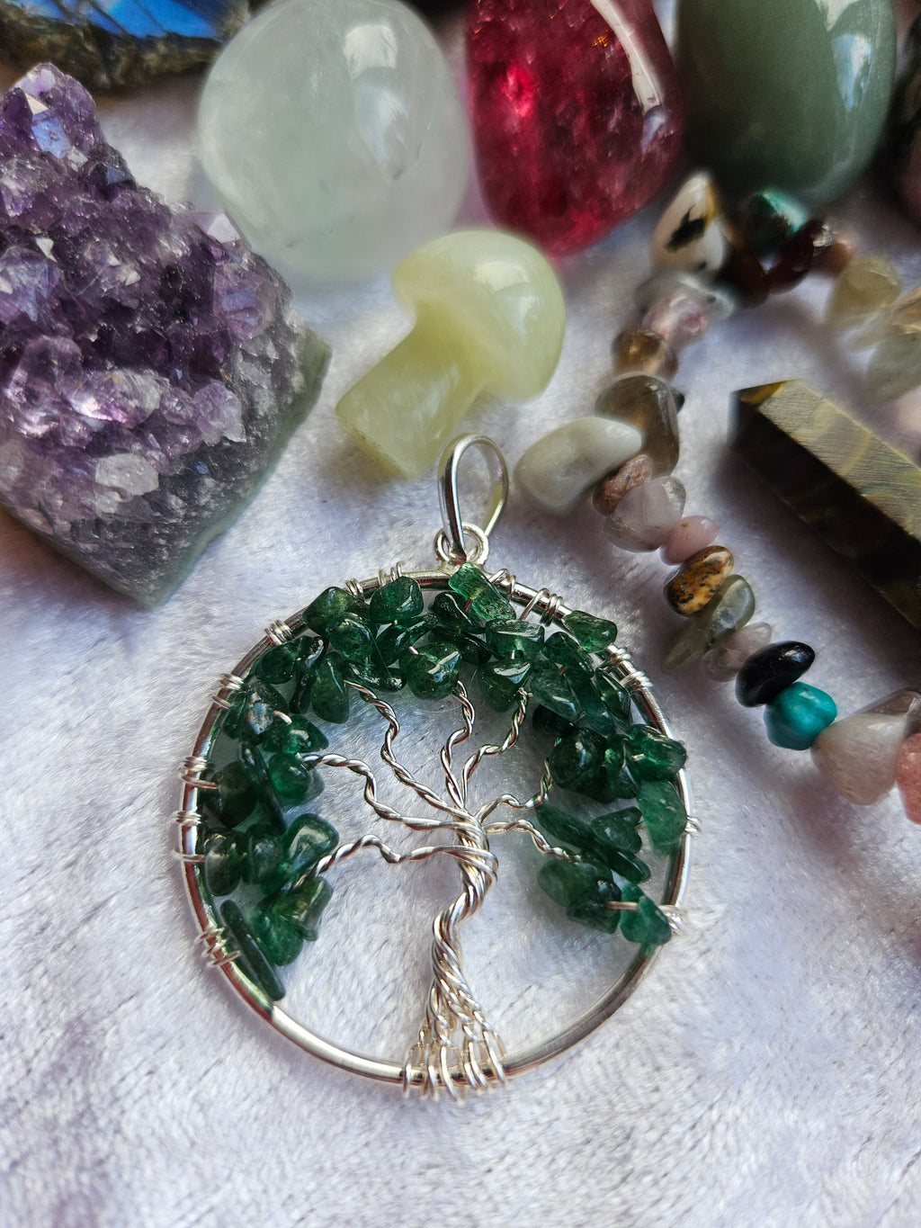 A silver wire-wrapped tree of life pendant with green gemstone leaves, displayed on a white textured surface surrounded by various colorful crystals and beads.
