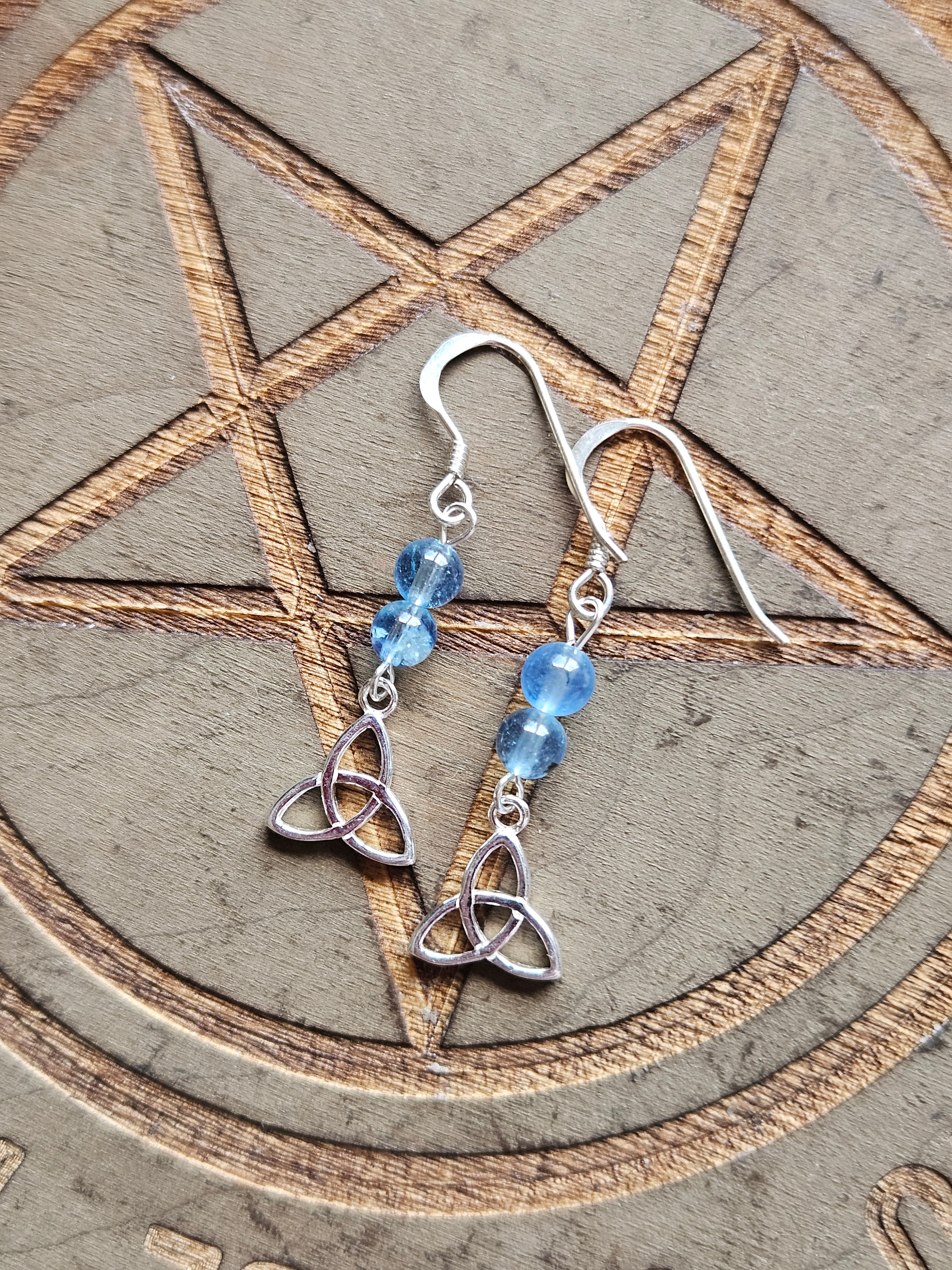 A pair of Sterling Silver dangle earrings with blue Aquamarine beads and a Celtic Triskele knot charm, displayed on a textured surface with a pentagram design.