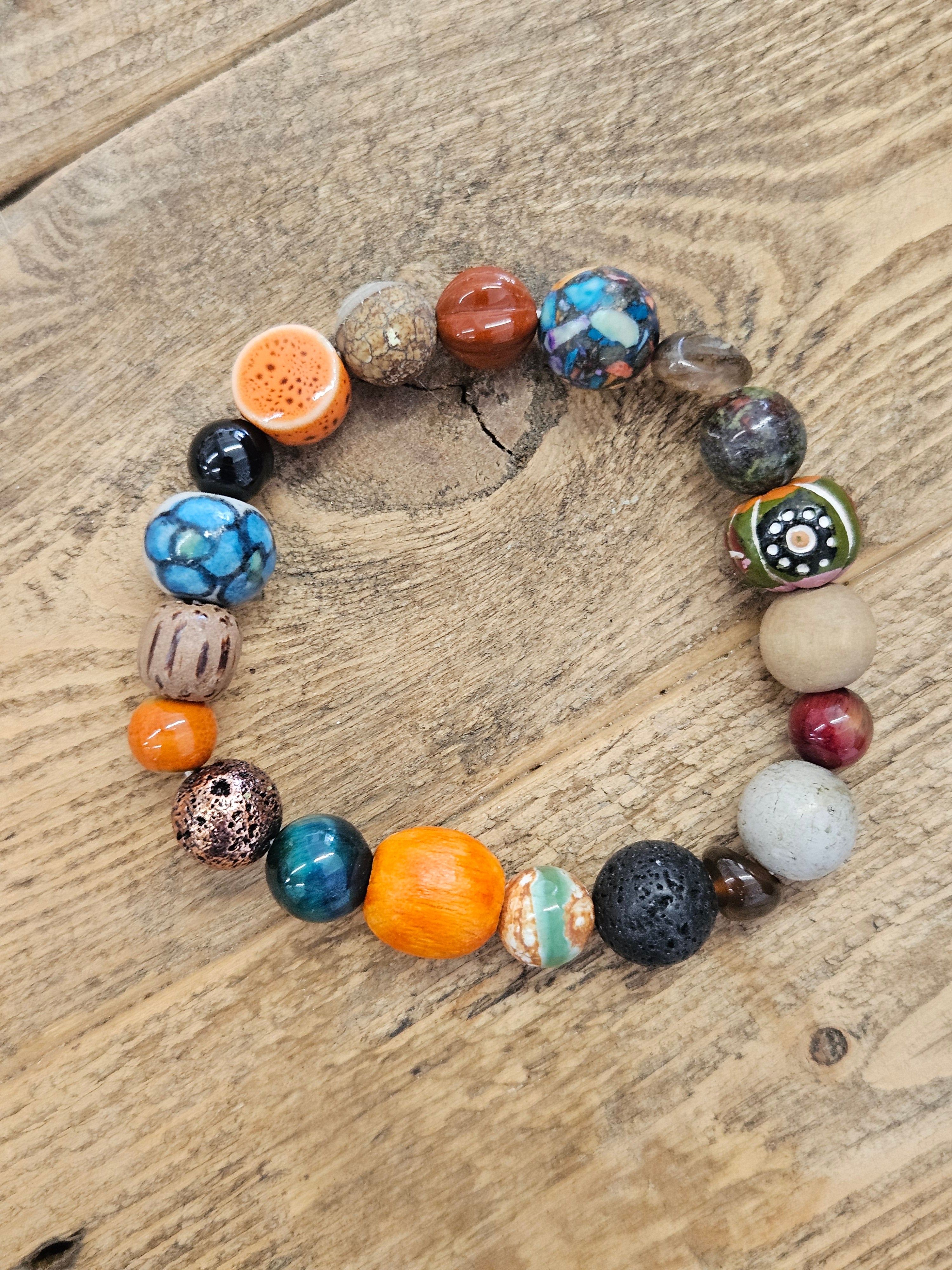 A handmade beaded stretch bracelet arranged in a circle on a rustic wooden surface. The bracelet features a variety of colorful and patterned beads, including orange, blue, black, brown, and white, with one bead shaped like a cross-section of an orange and another with a geometric dot pattern.