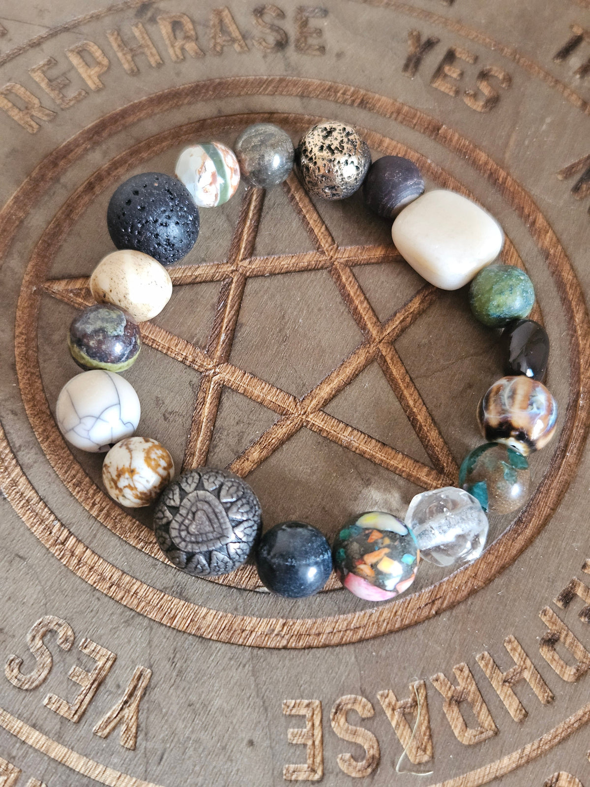 A beaded boho bracelet arranged in a circle on a wooden pentagram and altar board. The beads are made of various natural stones and crystals in multiple colors, including black, white, green, brown, and metallic gold.